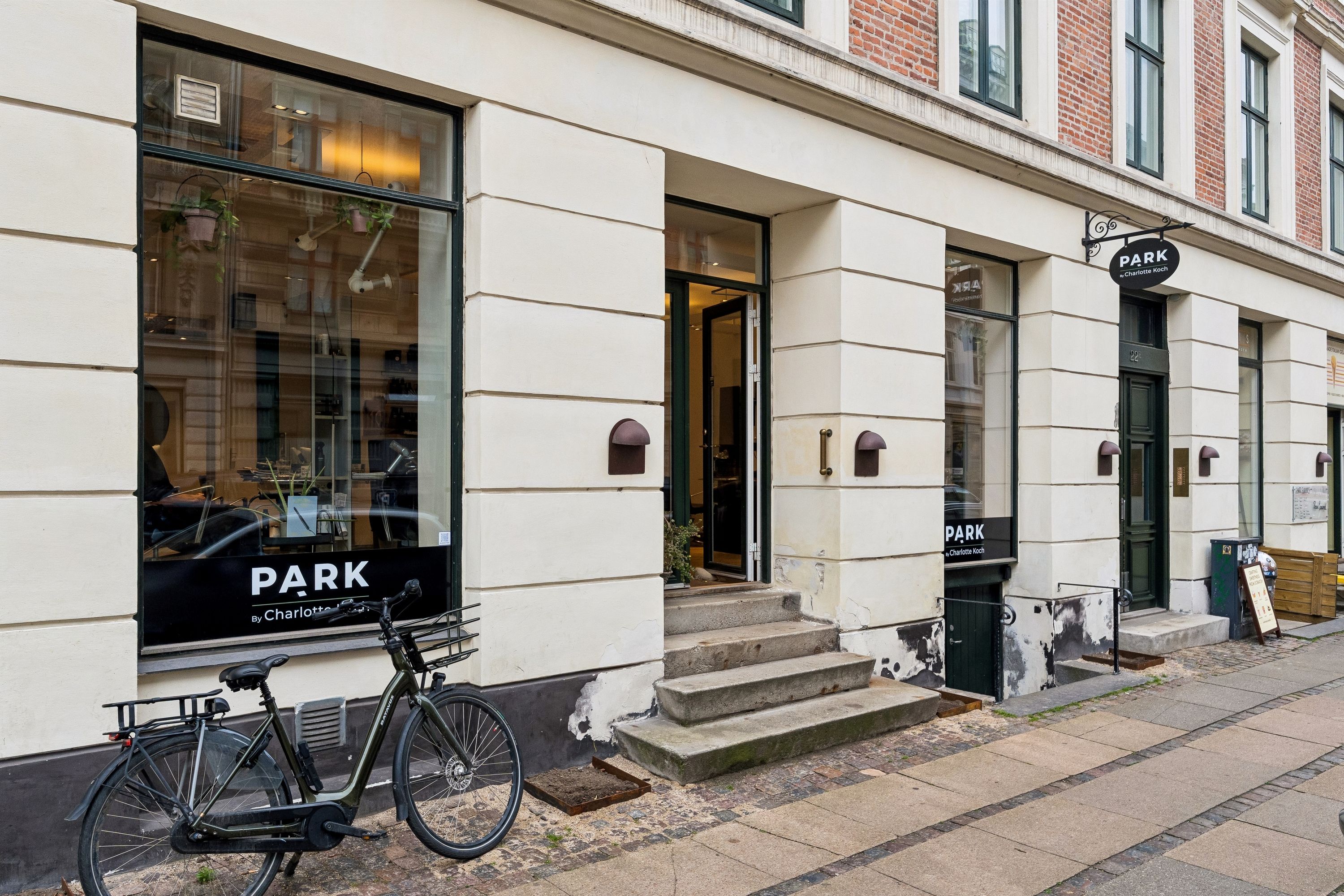 Frisørsalonen Park Charlotte Koch ligger på Holbergsgade i København. Stedets facade er malet hvidt og der er grønne plantekrukker. Der er også en cykel parkeret foran døren