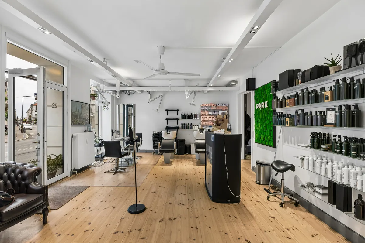 Elegant frisørsalon med moderne interiør og højt loft. Højrekanten er prydet med et grønt vægkunstværk og en skænk med flere produkter. Salonen har to højstole og flere barberstole i midten