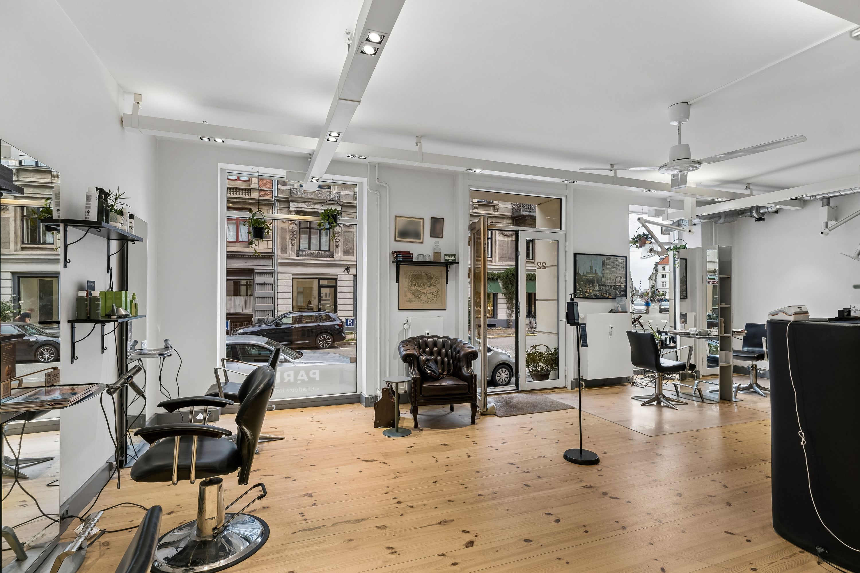 Moderne frisørsalon med lyse lofter og store glasdøre. Indenfor er der barberstole og lædersofaer samt et lille bord med en plante. Loftventilatorer og lysekroner øverst.