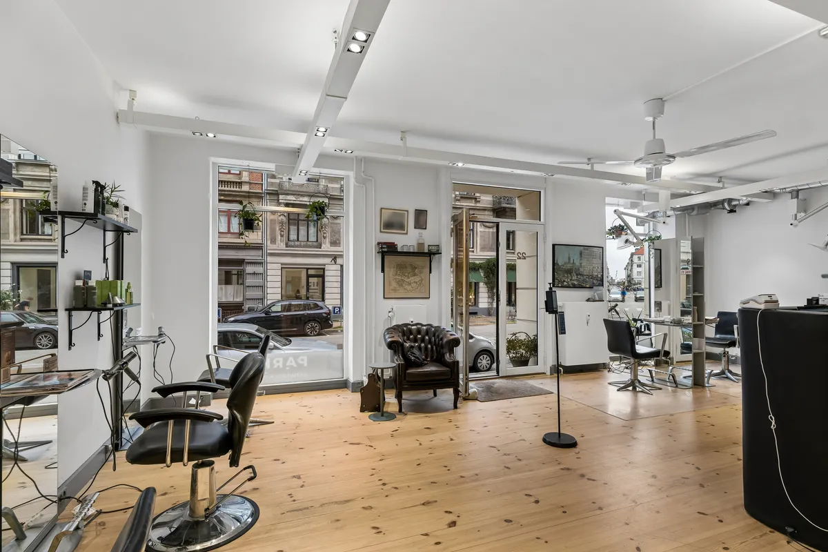 Moderne frisørsalon med lyse lofter og store glasdøre. Indenfor er der barberstole og lædersofaer samt et lille bord med en plante. Loftventilatorer og lysekroner øverst.