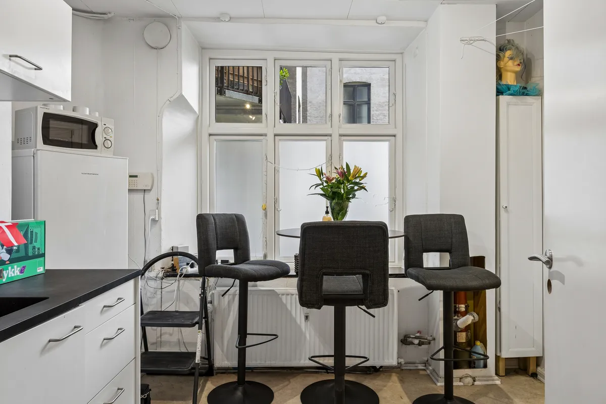 En hvid køkken med en hvid mikrobølgeovn og køleskab og en skab med en dukkehoved. Tre høje stole med lyserøde blomster i et glas og en hvid radiator under vinduet
