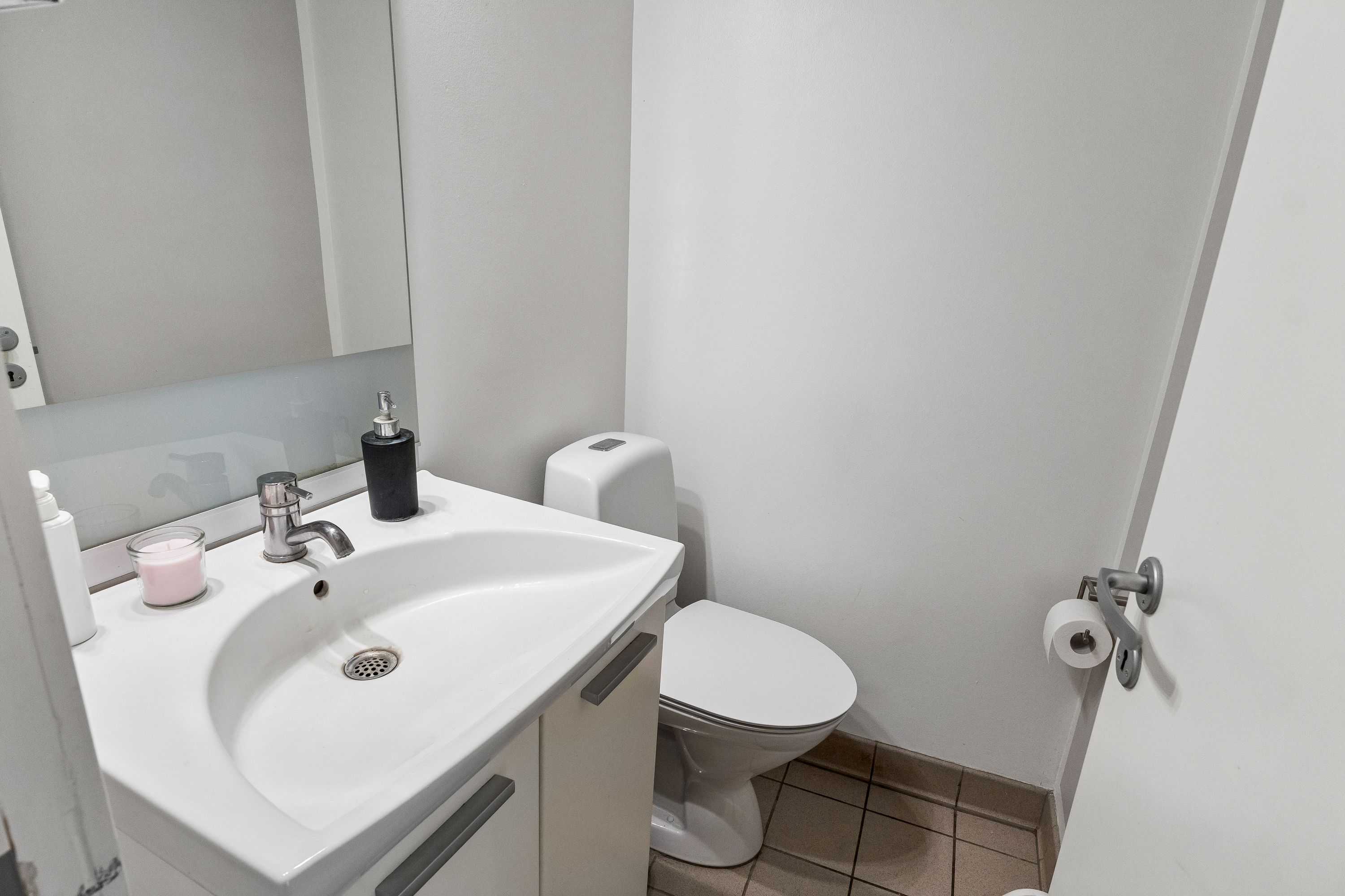 Et lille toiletrum med en hvid toilet, en hvid vaskeskål, håndvask med vandhaner og en toiletbørste og et toiletpapirhylde med to ruller toiletpapir og en hånddesinfektionsflaske