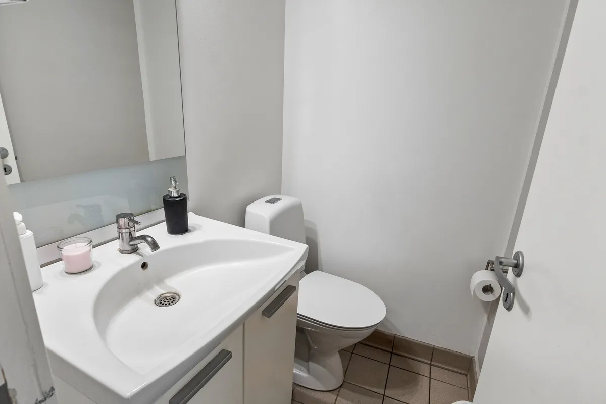 Et lille toiletrum med en hvid toilet, en hvid vaskeskål, håndvask med vandhaner og en toiletbørste og et toiletpapirhylde med to ruller toiletpapir og en hånddesinfektionsflaske
