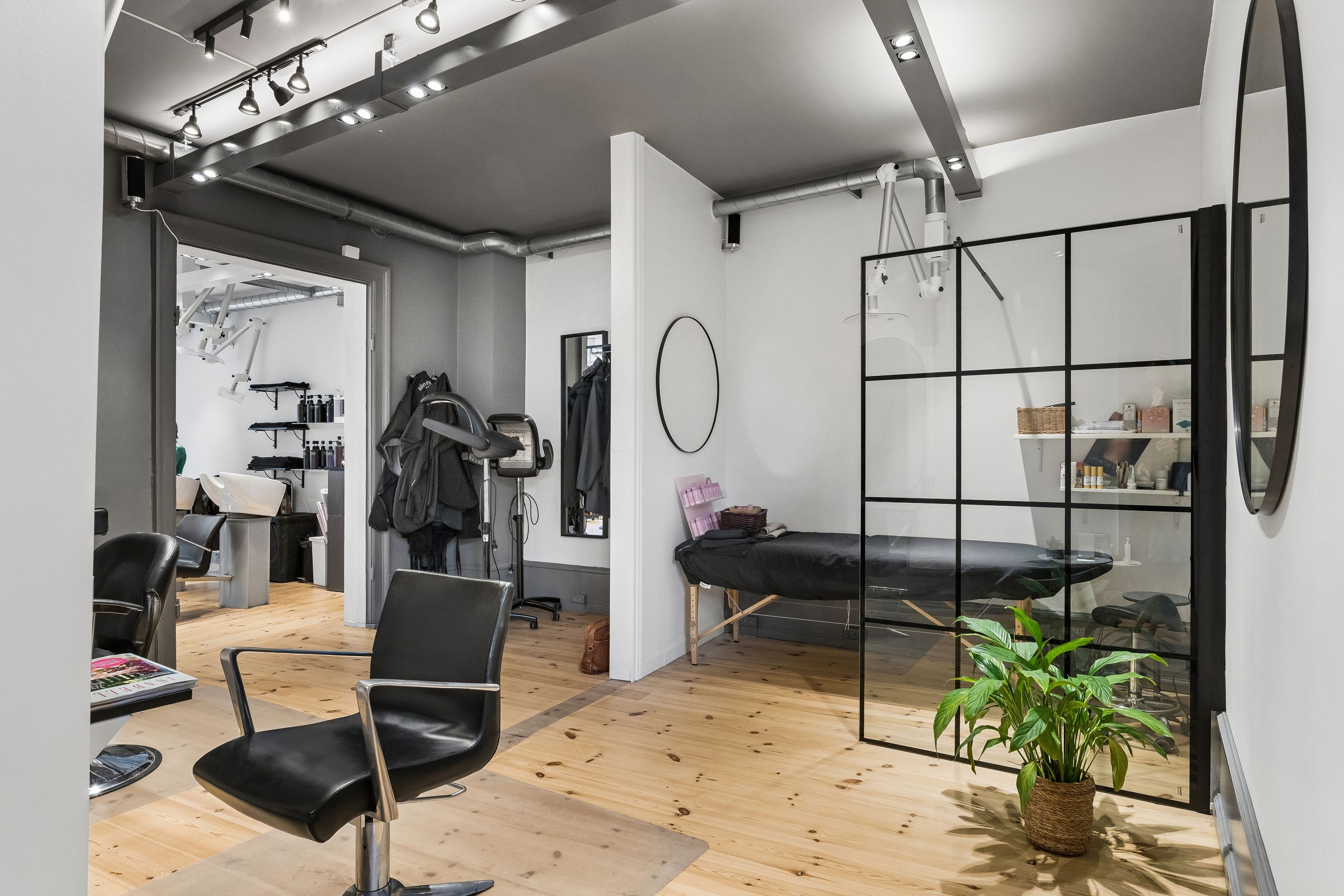 Inde i en moderne frisørsalon med lyse gulve og hvide vægge er der en stol, et spejl og en plante i en kurv ved vinduet