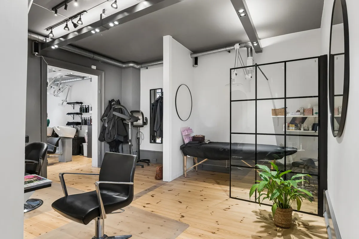 Inde i en moderne frisørsalon med lyse gulve og hvide vægge er der en stol, et spejl og en plante i en kurv ved vinduet