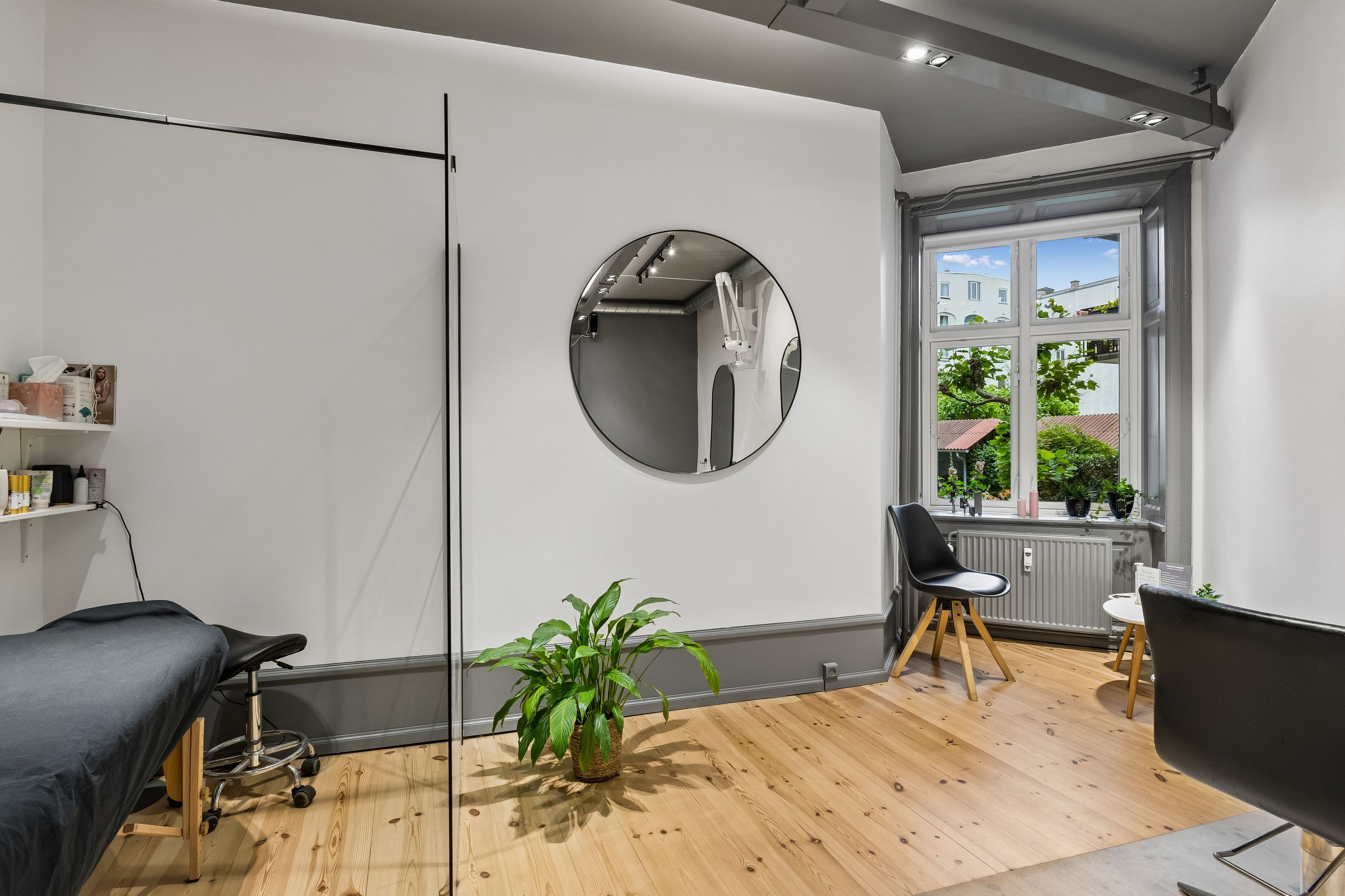 Dansk frisørsalon med runde spejl, højt loft, og en plante i krukken, samt et enkelt stol og et vindue med udsigt til byen