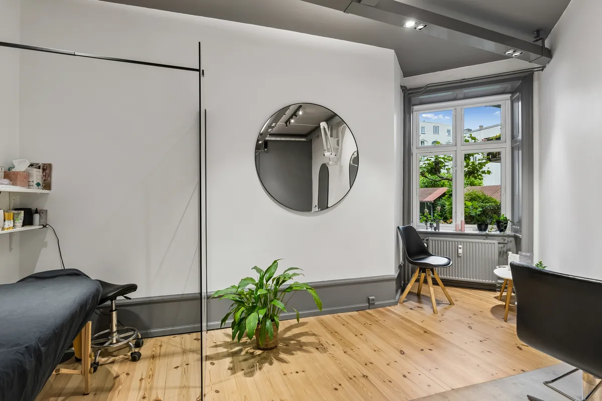 Dansk frisørsalon med runde spejl, højt loft, og en plante i krukken, samt et enkelt stol og et vindue med udsigt til byen