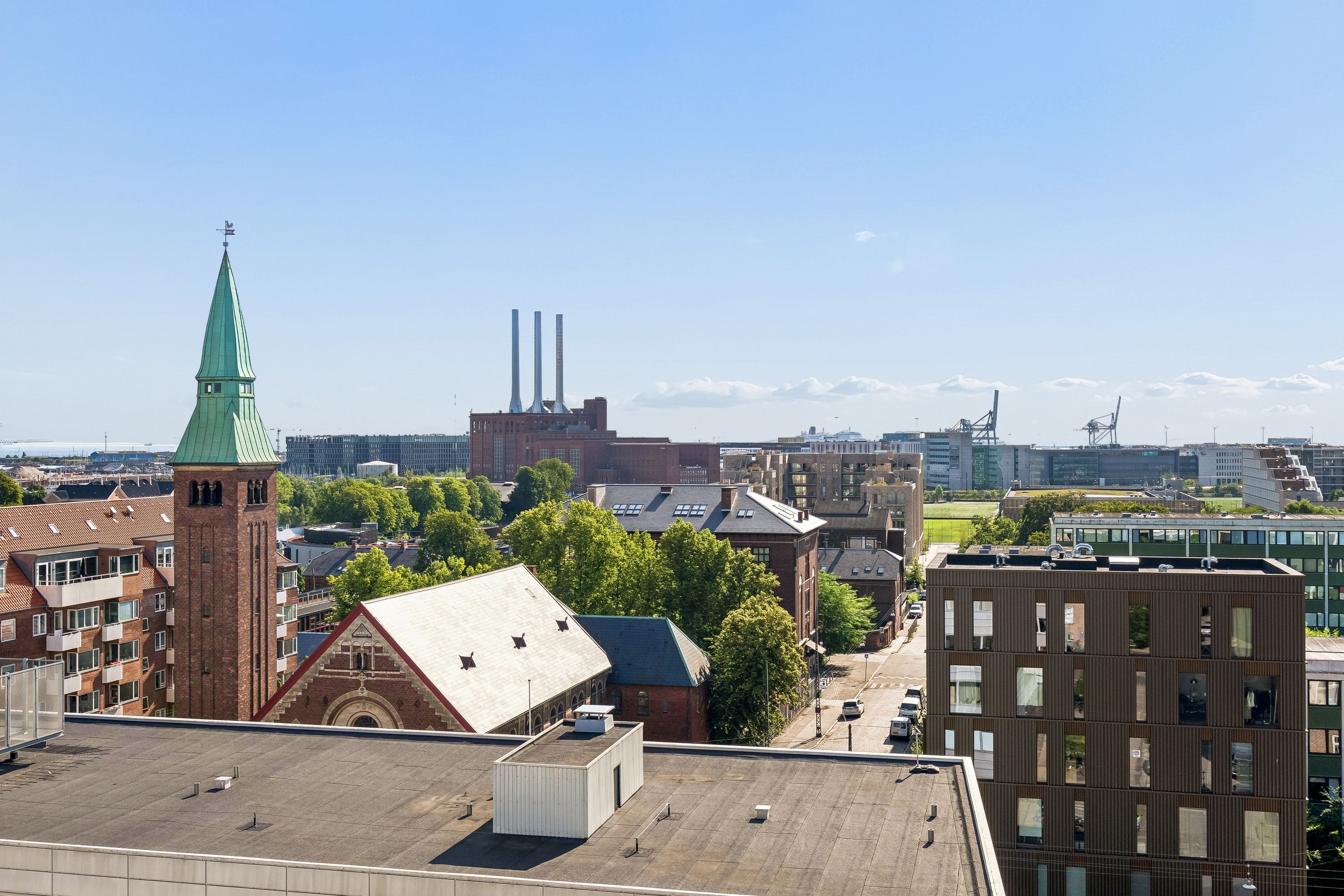 Lyst og indbydende kontorlejemål på Østerbrogade med udsigt over København. Bygningen med røde træk og grøn tagkant. I baggrunden er fabriksbygninger og søhavn.