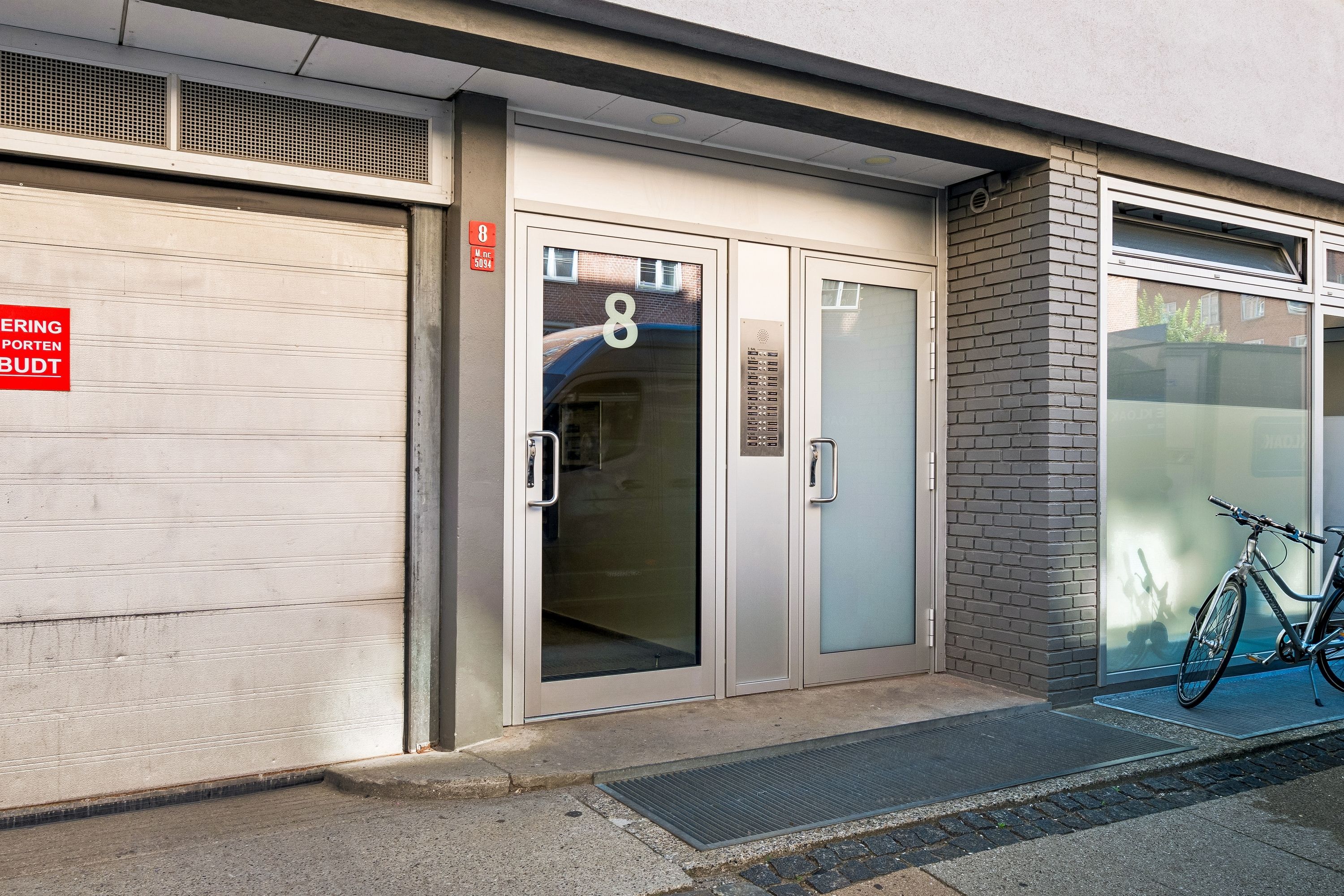 En moderne bygning med glasdøre og skilt med nummer 8. Cykel står parkeret med nummer 8. En garagedør udenfor. Solskin skinner på glasdøre og gulve
