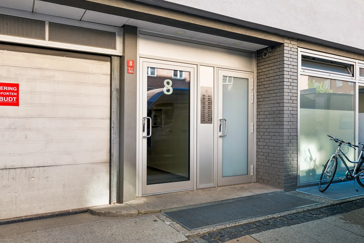 En moderne bygning med glasdøre og skilt med nummer 8. Cykel står parkeret med nummer 8. En garagedør udenfor. Solskin skinner på glasdøre og gulve