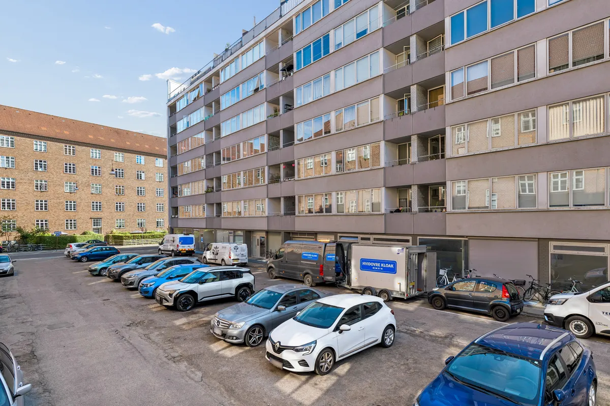Flere biler parkeret på en parkeringsplads med en stor bygning bagved og flere vinduer og balkoner