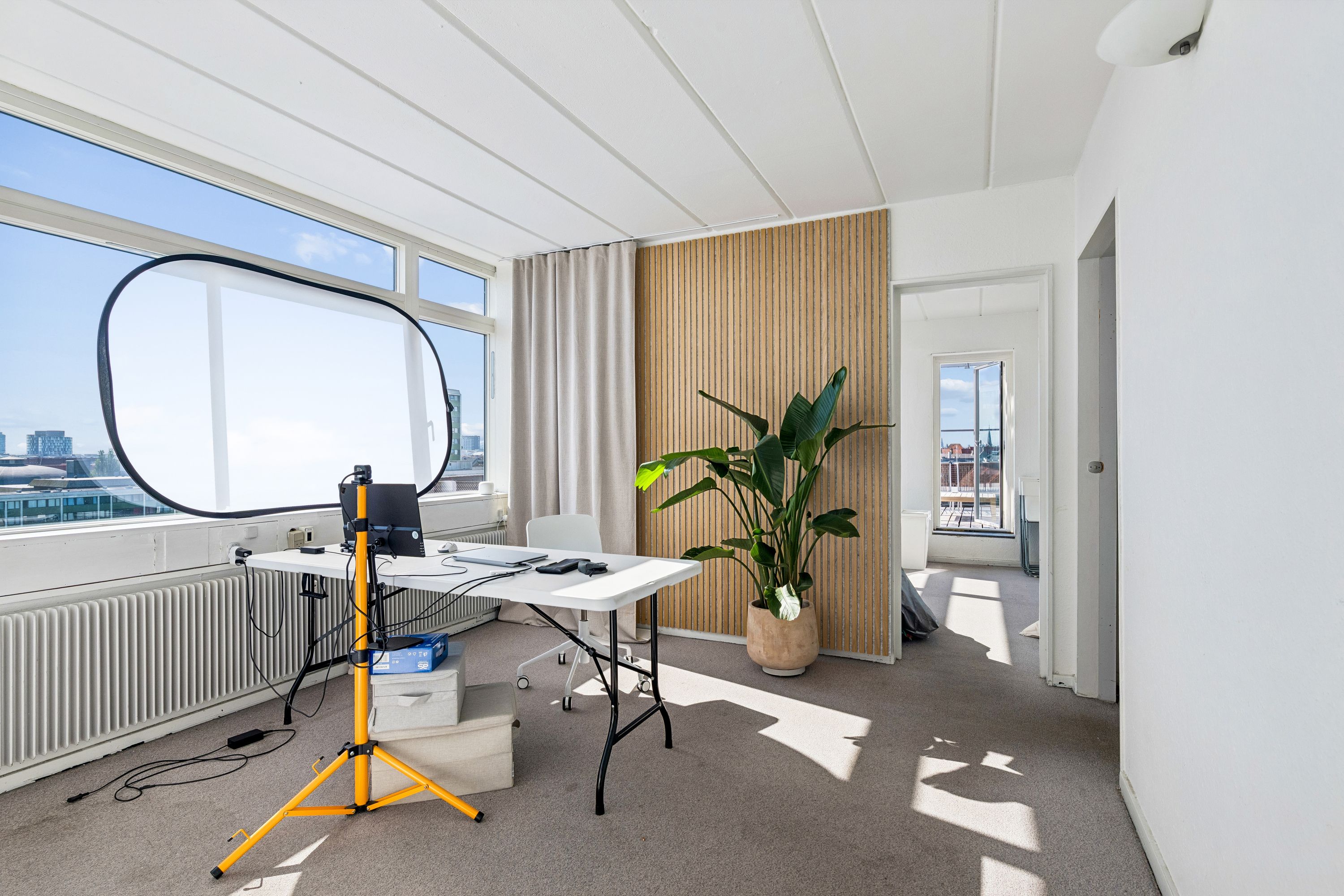 Kontor med hvidt bord, stålampe, plante, stol og kamera. Vindue med udsigt over byen. Solskin på gulvet.