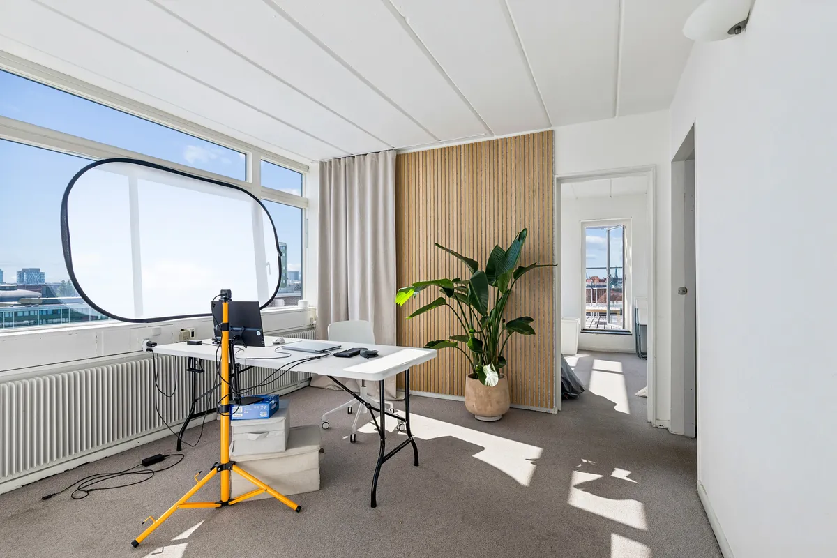 Kontor med hvidt bord, stålampe, plante, stol og kamera. Vindue med udsigt over byen. Solskin på gulvet.