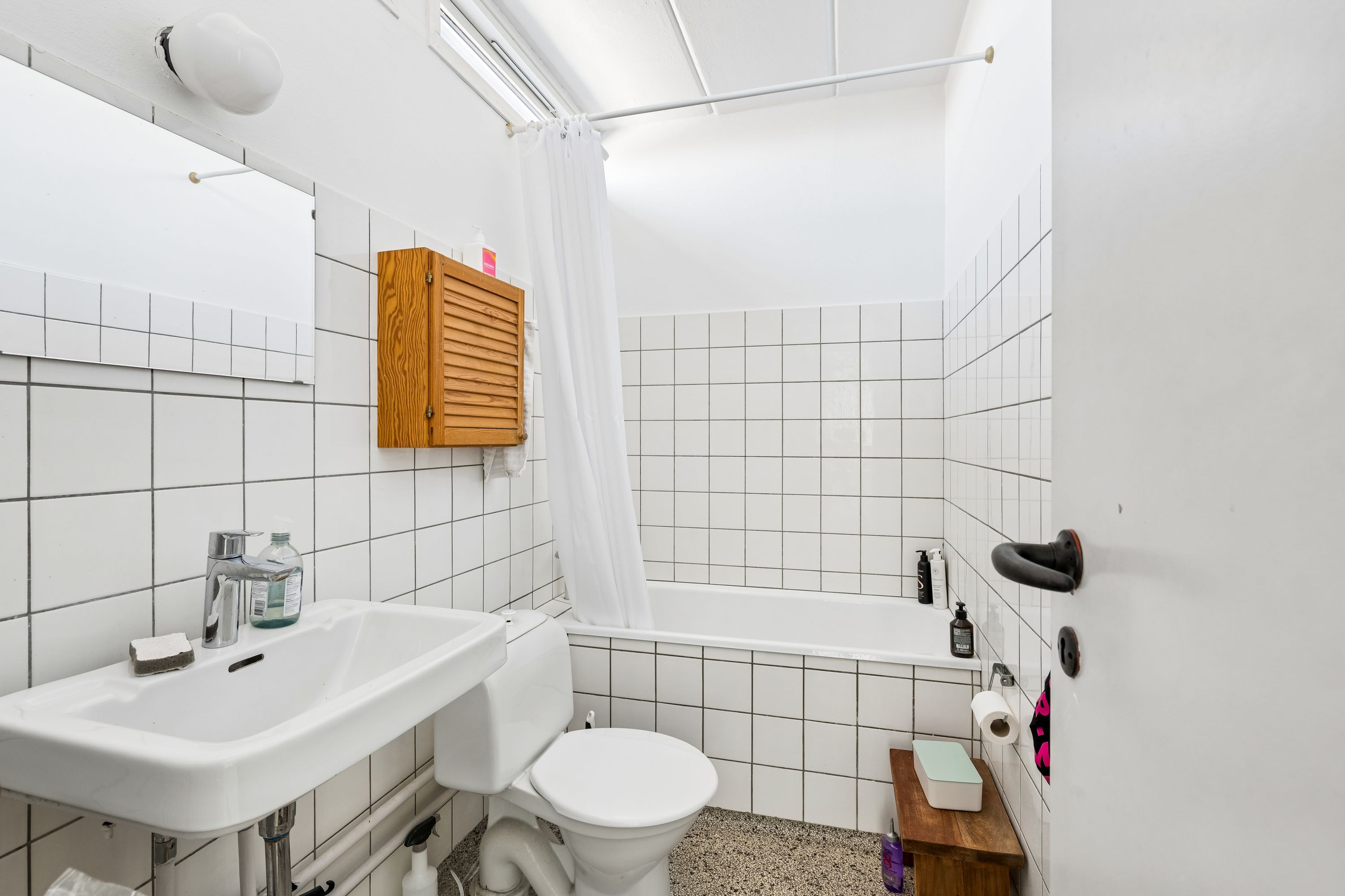 Hvidt badeværelse med hvidt toilet, hvidt badekar, hvidt håndvask, hvidt håndklæde og tøjtørrer samt håndklæde og toiletpapir på toiletbordet