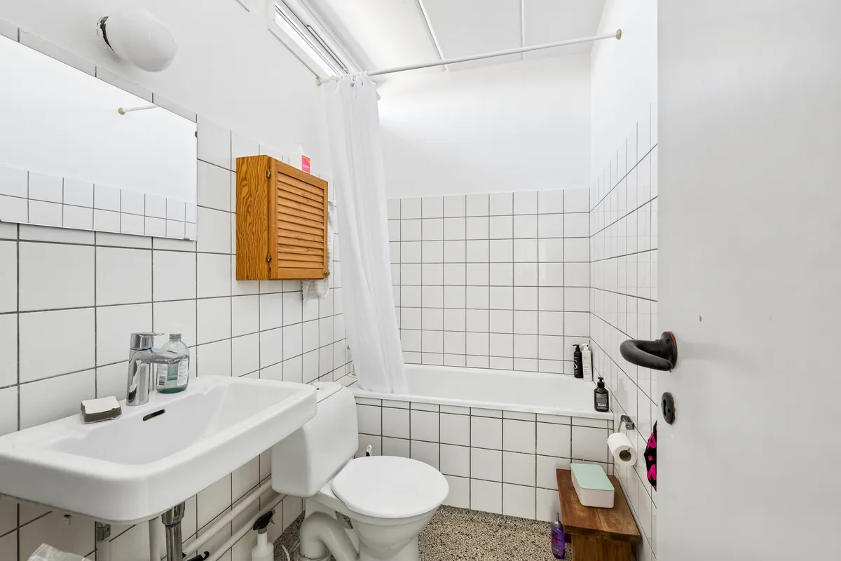 Hvidt badeværelse med hvidt toilet, hvidt badekar, hvidt håndvask, hvidt håndklæde og tøjtørrer samt håndklæde og toiletpapir på toiletbordet