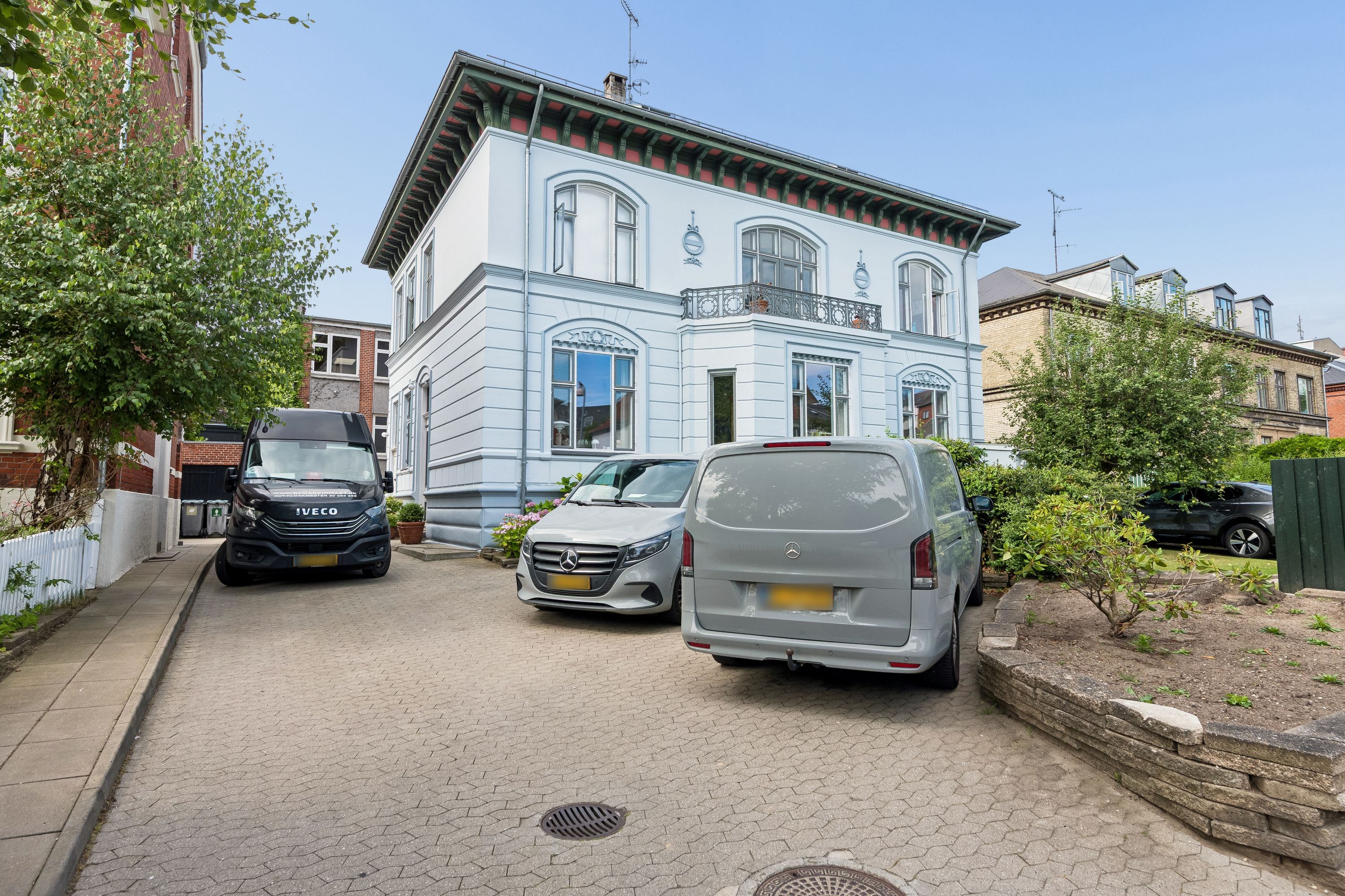 To personbiler og en varevogn parkeret uden for et kontorhuse med moderne facade og balkon på en solrig dag