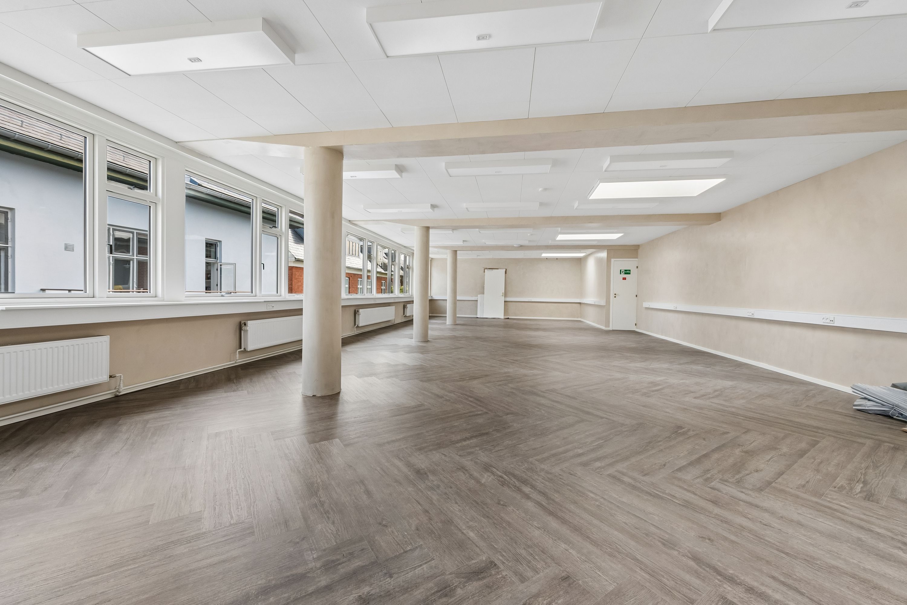 Kontor med moderne hvide lofter og store vinduer, trægulv og hvide vægge, radiatorer og to døre i baggrunden, sammen med lyse og funktionelle kontorråd.