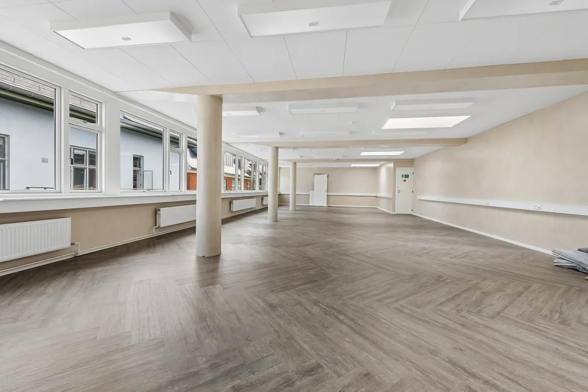 Kontor med moderne hvide lofter og store vinduer, trægulv og hvide vægge, radiatorer og to døre i baggrunden, sammen med lyse og funktionelle kontorråd.