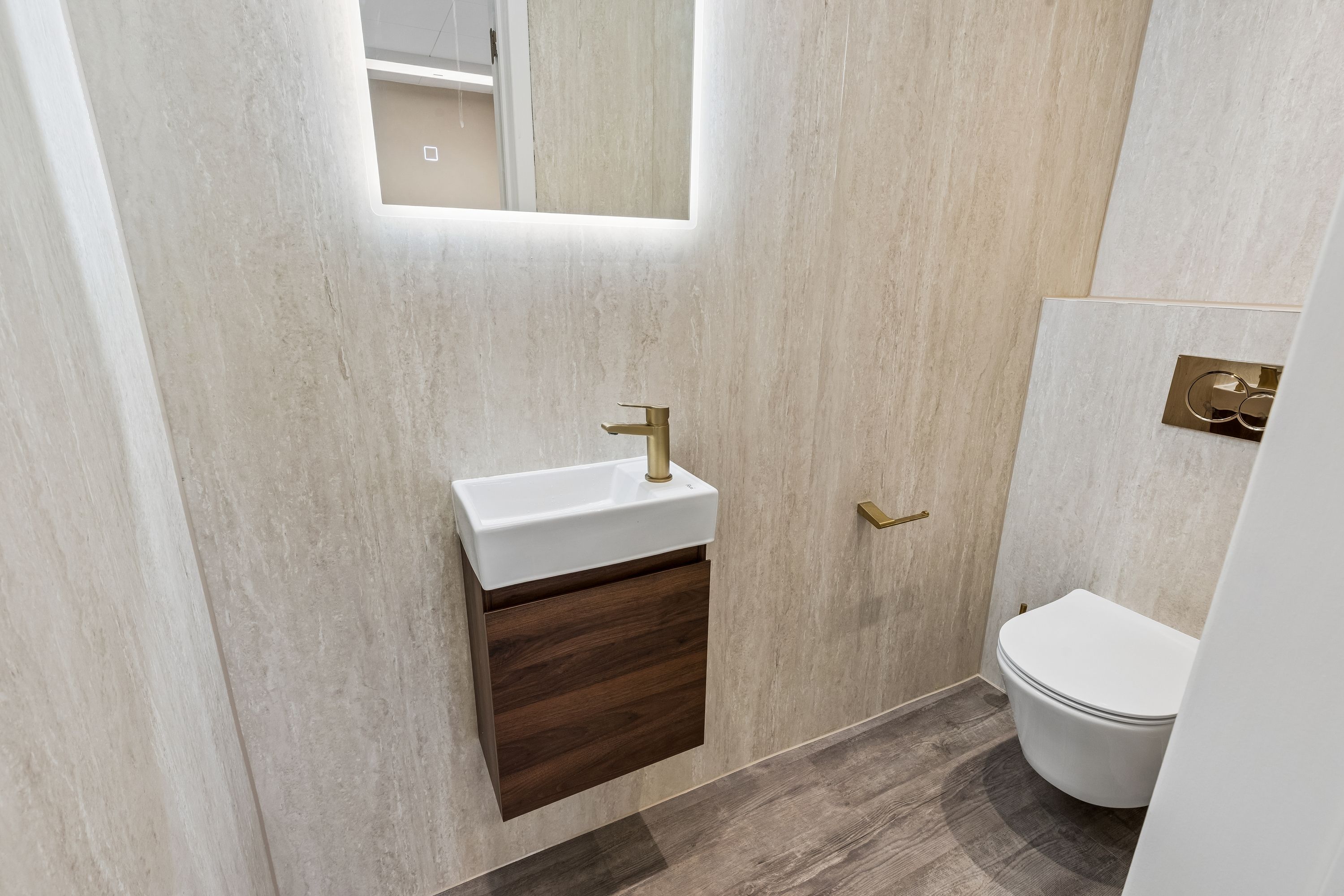 Nyrenoveret toilet med en marmorfarvet væg og en hvid toilet. Et højt gulv med trælignende møbler og en hvid marmorvask med en messingfarvet vandhane