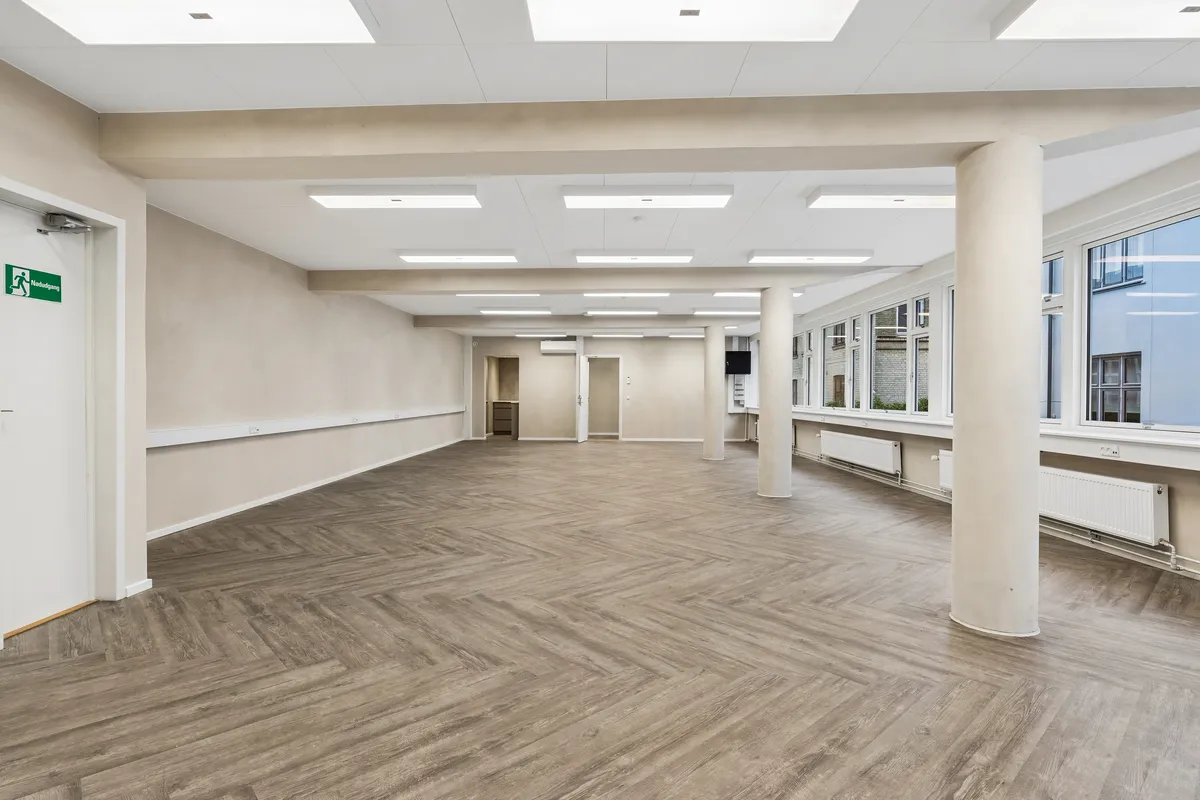 Ledig og rummelig kontor i Frederiksberg med lyse loftsbærende søjler, moderne gulvbelægning, hvide vægge og store vinduer til fuld solbelysning. Der er tekniske installationer og radiatorer langs væggene
