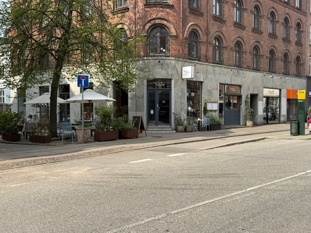 Take-away, bageri og deli på Gammel Kongevej. Sted med flere udendørsborde og parasoller. To store planter og stolpe med skilt. Bord med hvidt borddug og lyserødt serviet.