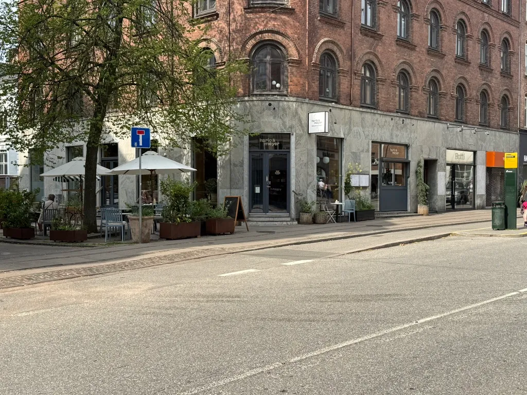 Take-away, bageri og deli på Gammel Kongevej. Sted med flere udendørsborde og parasoller. To store planter og stolpe med skilt. Bord med hvidt borddug og lyserødt serviet.