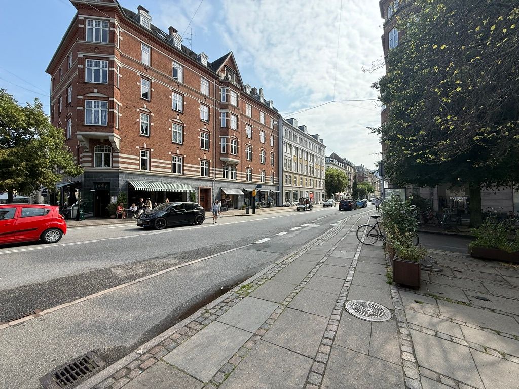 Gammel Kongevej med røde biler, hvidt og rødt bygningsfront, fortov, grønne planter, grønne træer og mennesker, der går på fortovet