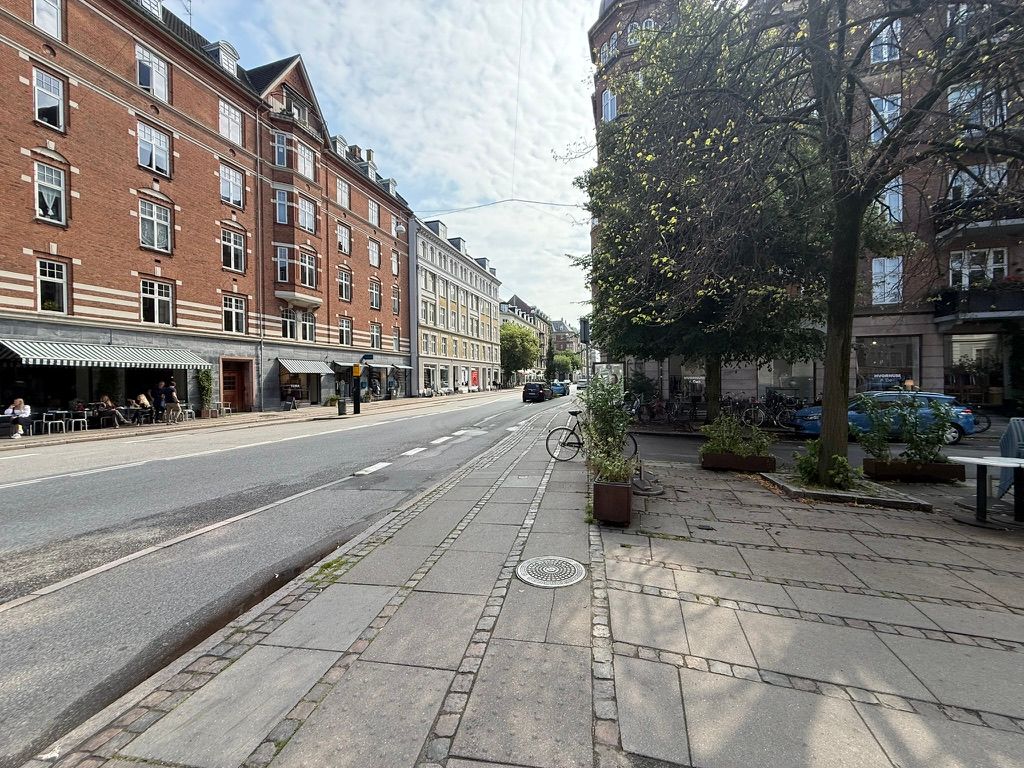 Take-away og bageri ligger på Gammel Kongevej 107. Både cykler og biler er parkeret langs gaden. Få mennesker sidder på terrassen med deres drikke og mad.