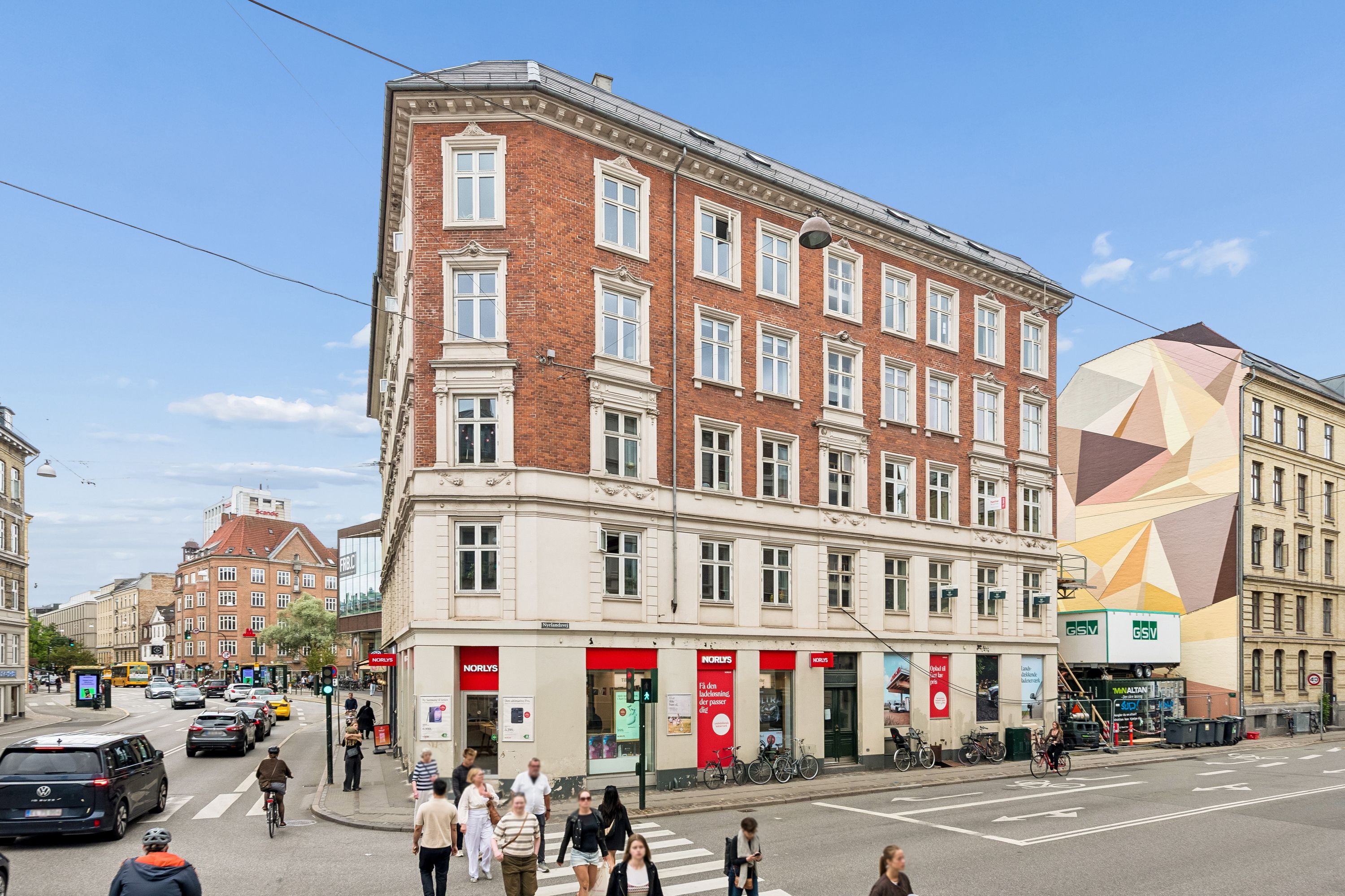 Mennesker går og cykler på gågaden med flere biler i baggrunden. Bygning med Norlys-butik og en grøn murmaleri på siden