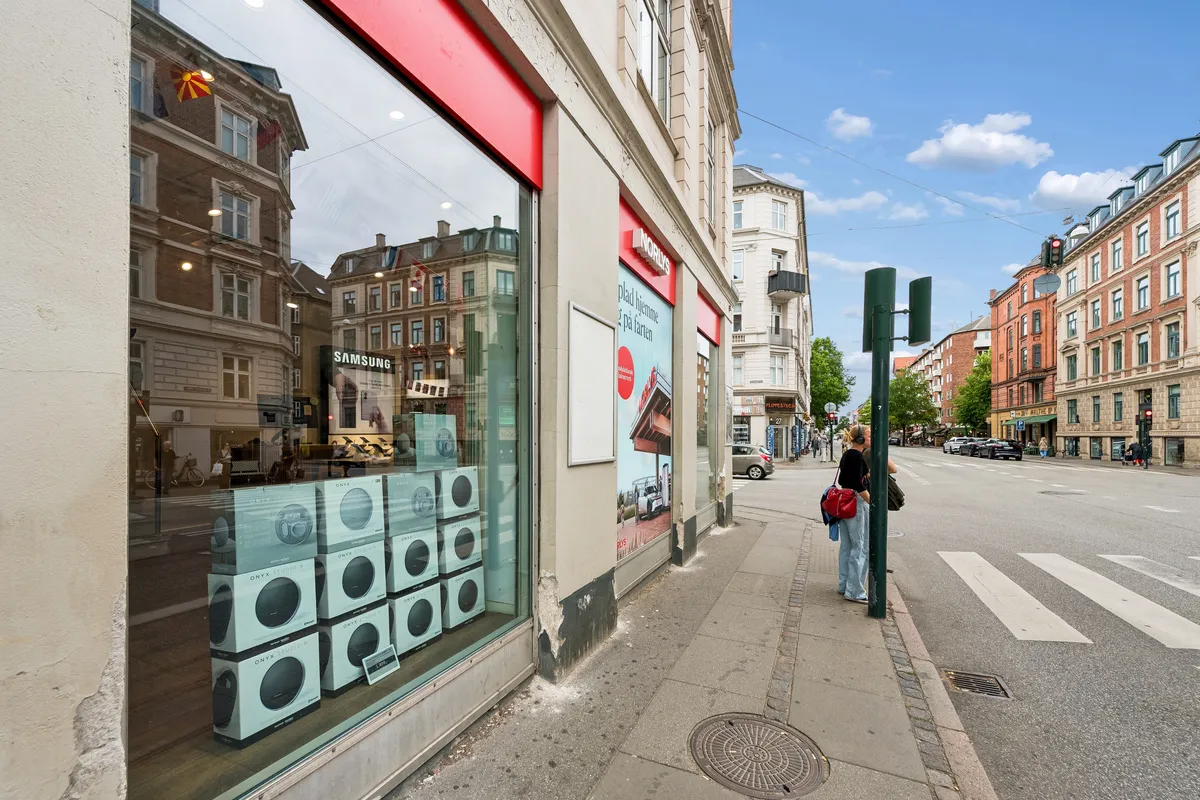 På Falkoner Alle står en butik med store vinduer og Samsung-udsalg. En kvinde venter på en gågade, og der er et stopsignal i baggrunden. Bygningen er klædt i beige murværk.