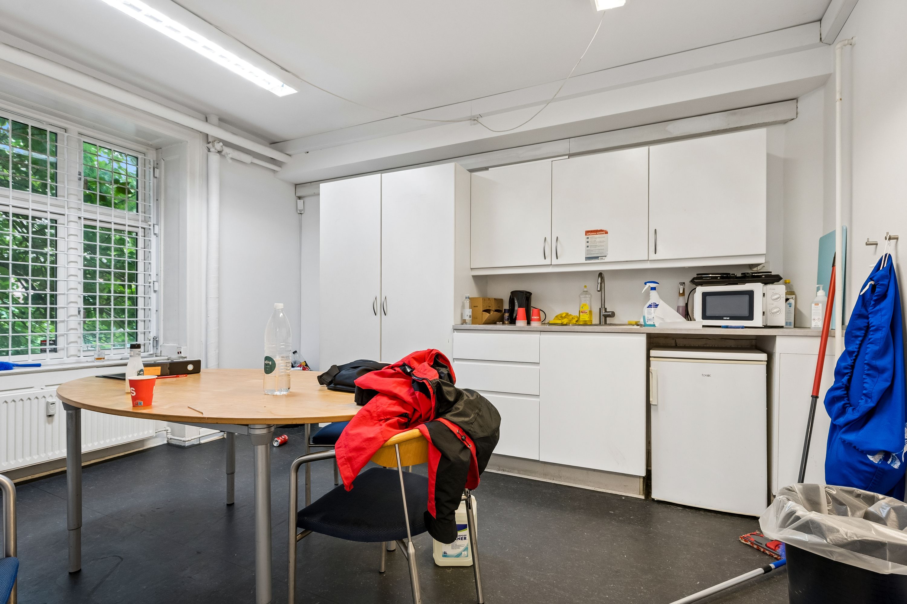 Rundt bord med rød plast kop og vandflaske. Hængende jakke og jakke på stol. Hvide hylder, mikrobølgeovn og køleskab. Renseriklud og støvsuger. Vindue med udsigt til grønne træer.