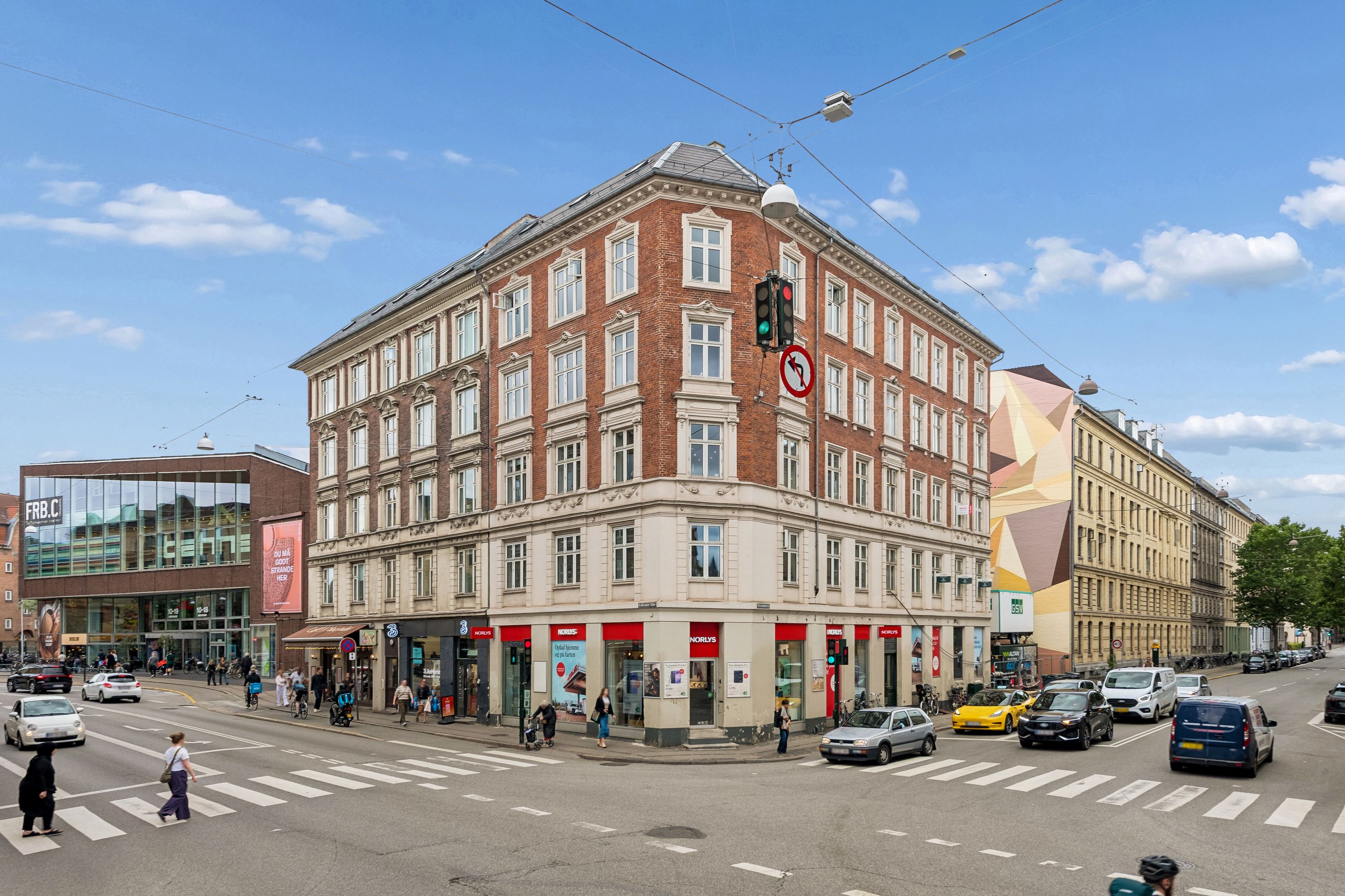 Hjørneejendom med butikslokaler og søjler på Falkoner Alle 25 i Frederiksberg med folk og biler, der passerer på vejen under en solrig dag