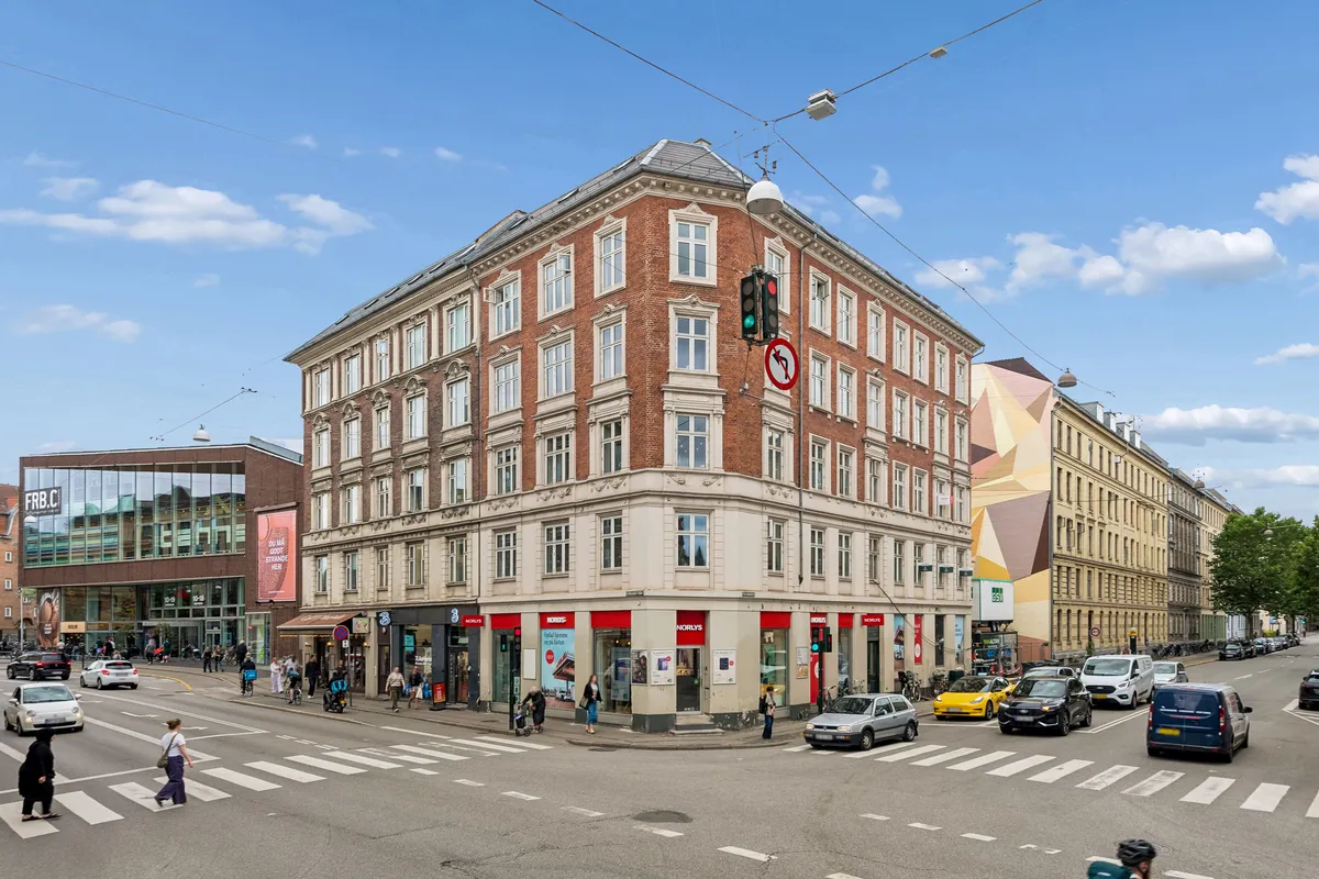 Hjørneejendom med butikslokaler og søjler på Falkoner Alle 25 i Frederiksberg med folk og biler, der passerer på vejen under en solrig dag