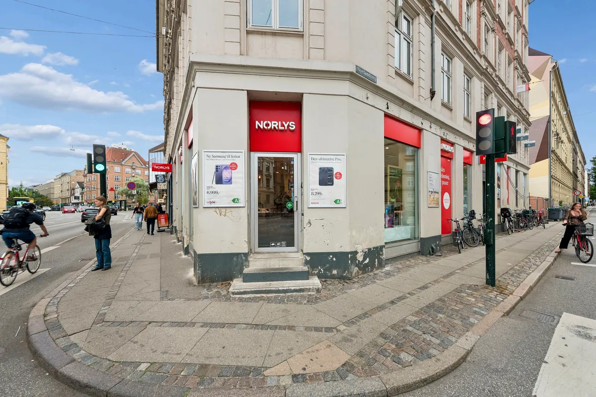 På Falkoner Alle 25, Frederiksberg, udbydes en 215 m² butikslokal til leje. Hjørneejendommen har Norlys-reklame og åbent indgangsdør. Flere cykler står parkeret og folk går på gågaden.