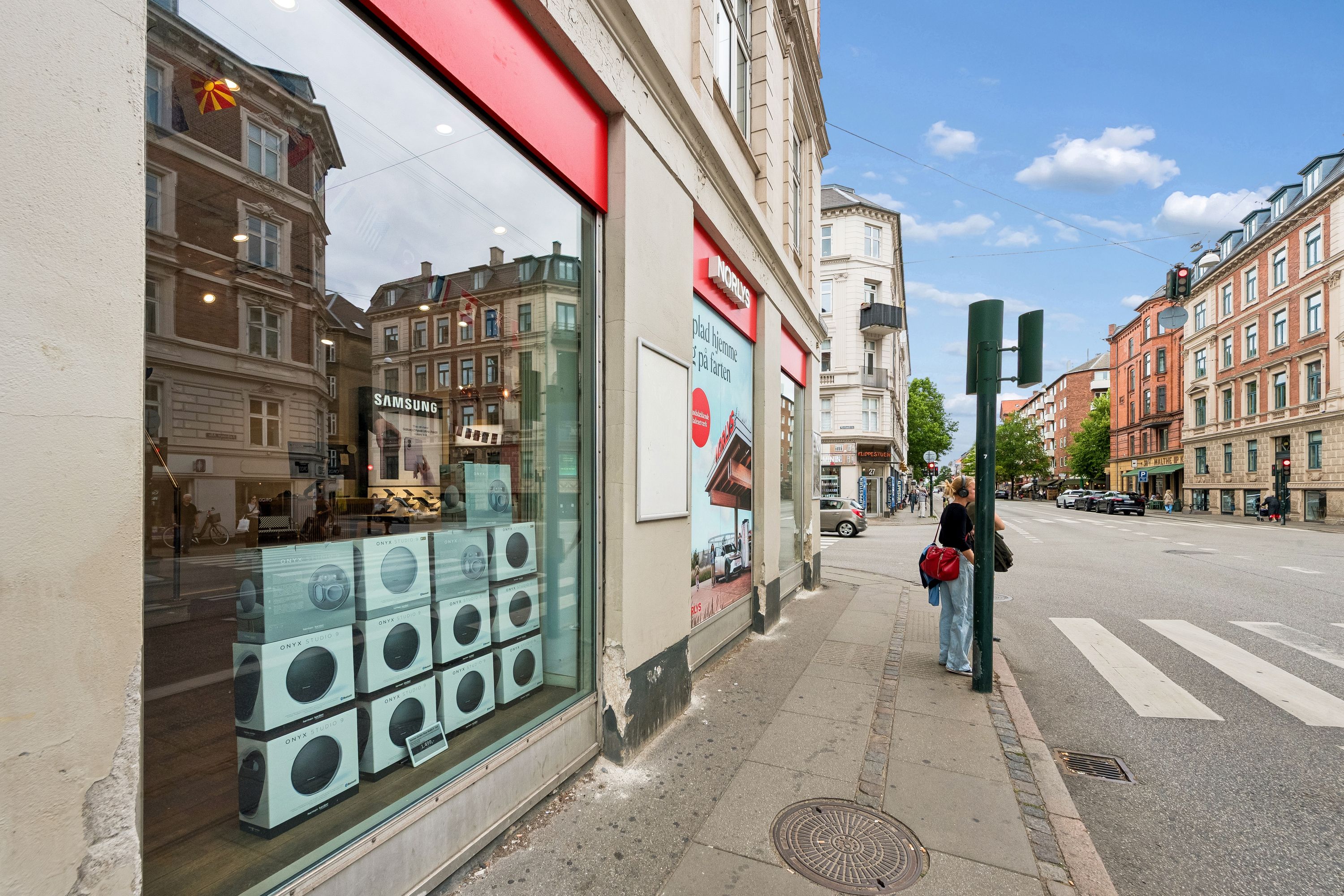 Butik med Samsung produkter og ONYX Studio 9 højttalere på Falkoner Alle i Frederiksberg. Bygningen med nordisk reklame og rødt hjørne. En kvinde står og læser avis på fortovet