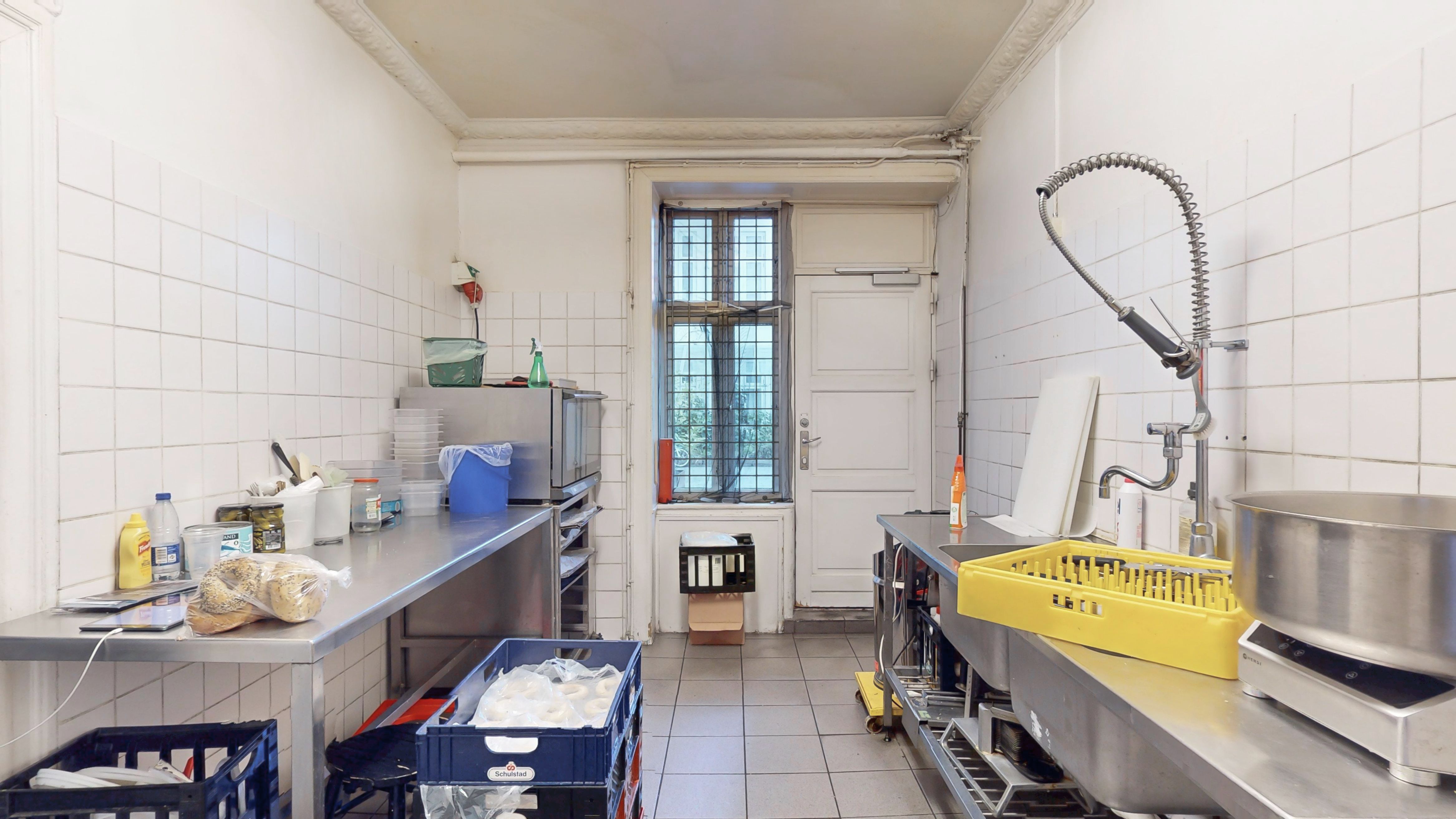 Lejet koldt køkken med bord, mikrobølgeovn, køleskab og flere opbevaringskurve. Skurvægge og gulv. Sink og stålkøkken med bøjler og stålfade.