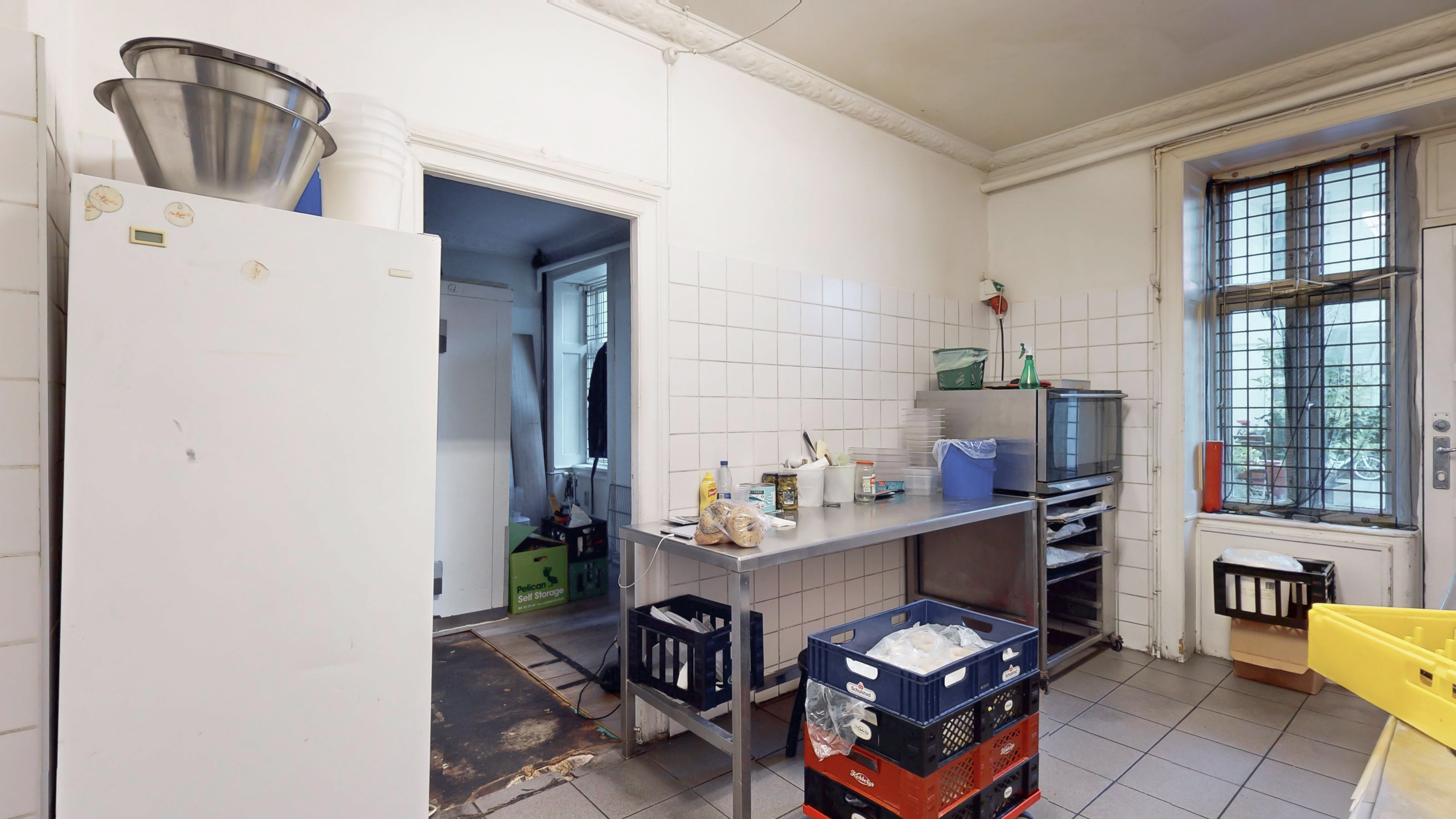 Koldt køkken med metalbord, aaben køleskab, metalregn og kassevogn med hjul i lysegråt værelse med hvidt klinkergulv og hvide vægge