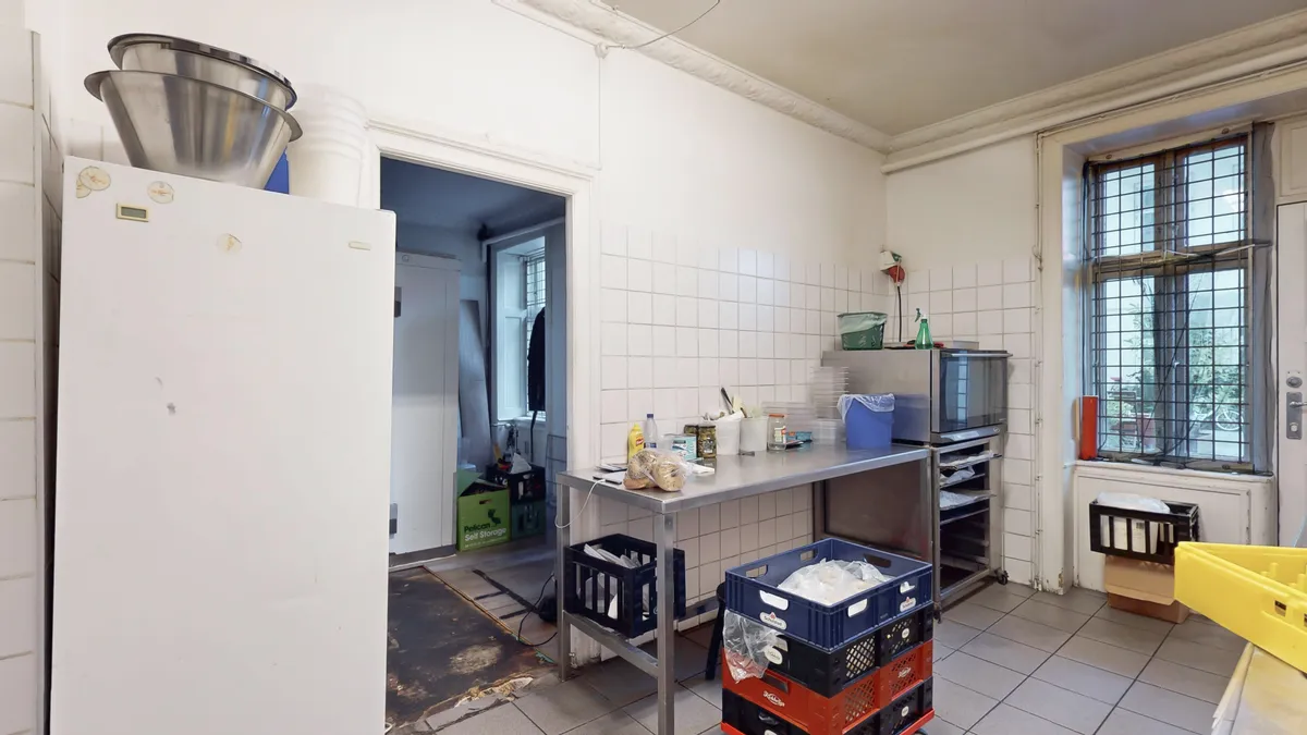 Koldt køkken med metalbord, aaben køleskab, metalregn og kassevogn med hjul i lysegråt værelse med hvidt klinkergulv og hvide vægge