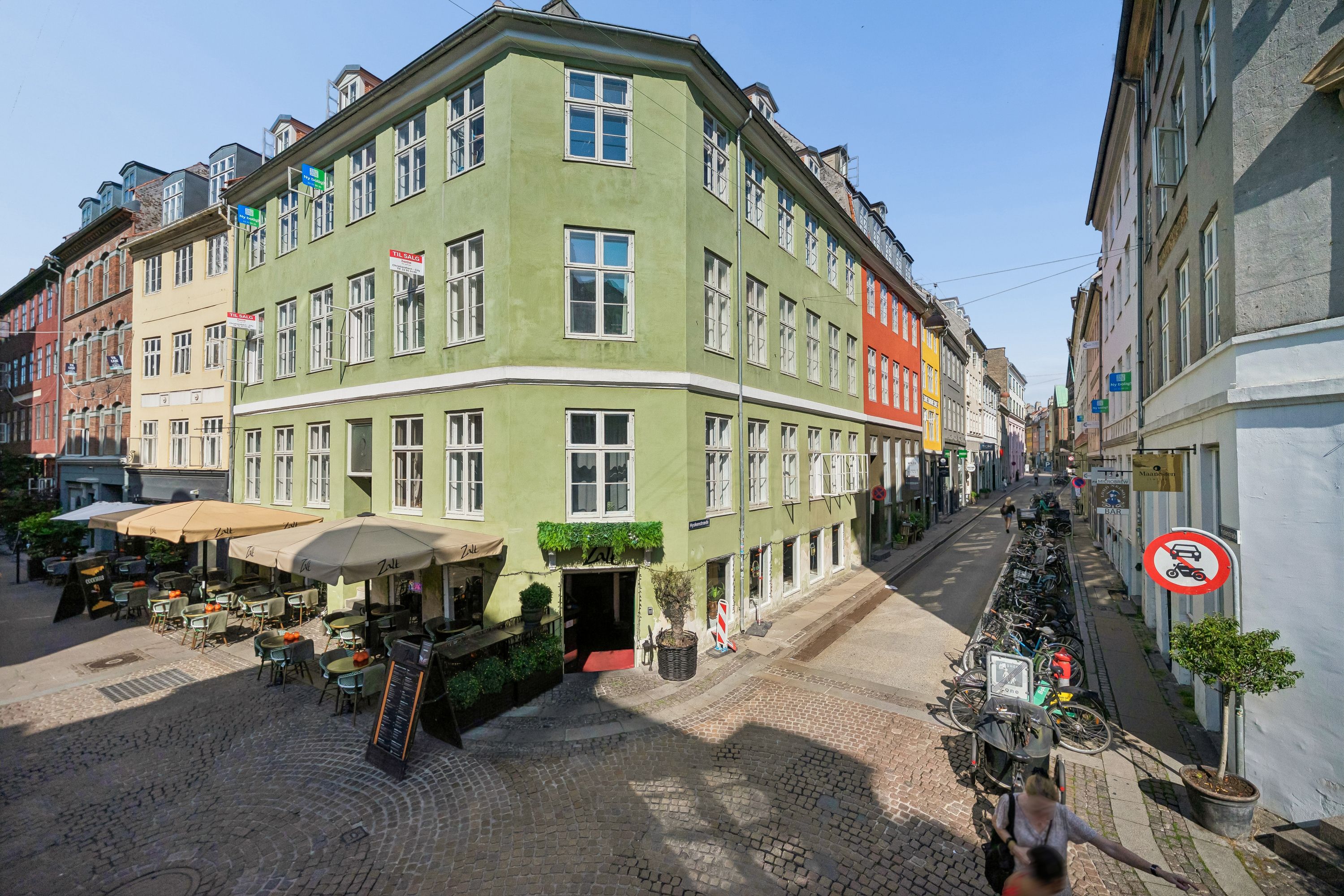 Baghuskontor på Hyskenstræde med grøn facade, solbeskyttelse, havemøbler og grønne plantager. Mange cykler og søjler langs den brolagte gade.