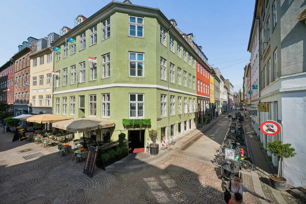 Baghuskontor på Hyskenstræde med grøn facade, solbeskyttelse, havemøbler og grønne plantager. Mange cykler og søjler langs den brolagte gade.