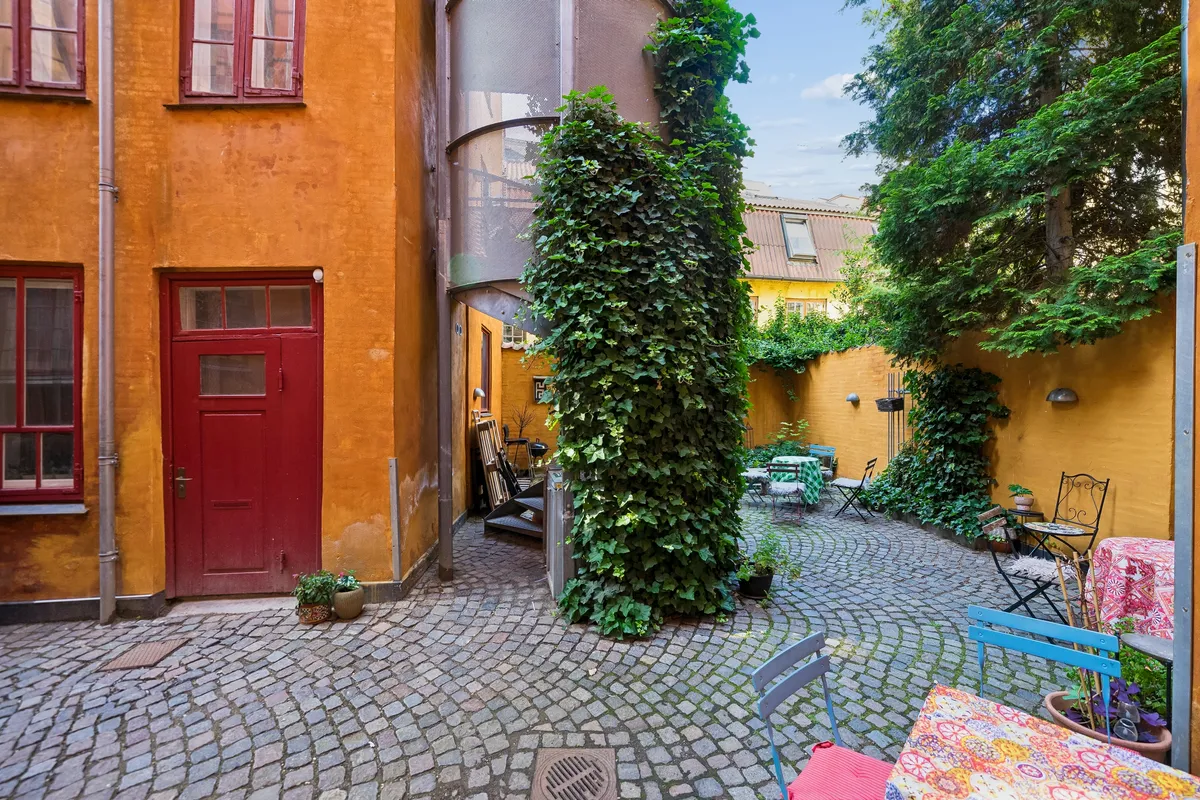 En lille gade med terrasse, stolpe og lyseplanter på væggen, gulvet er brolægget og der er en rødt dør