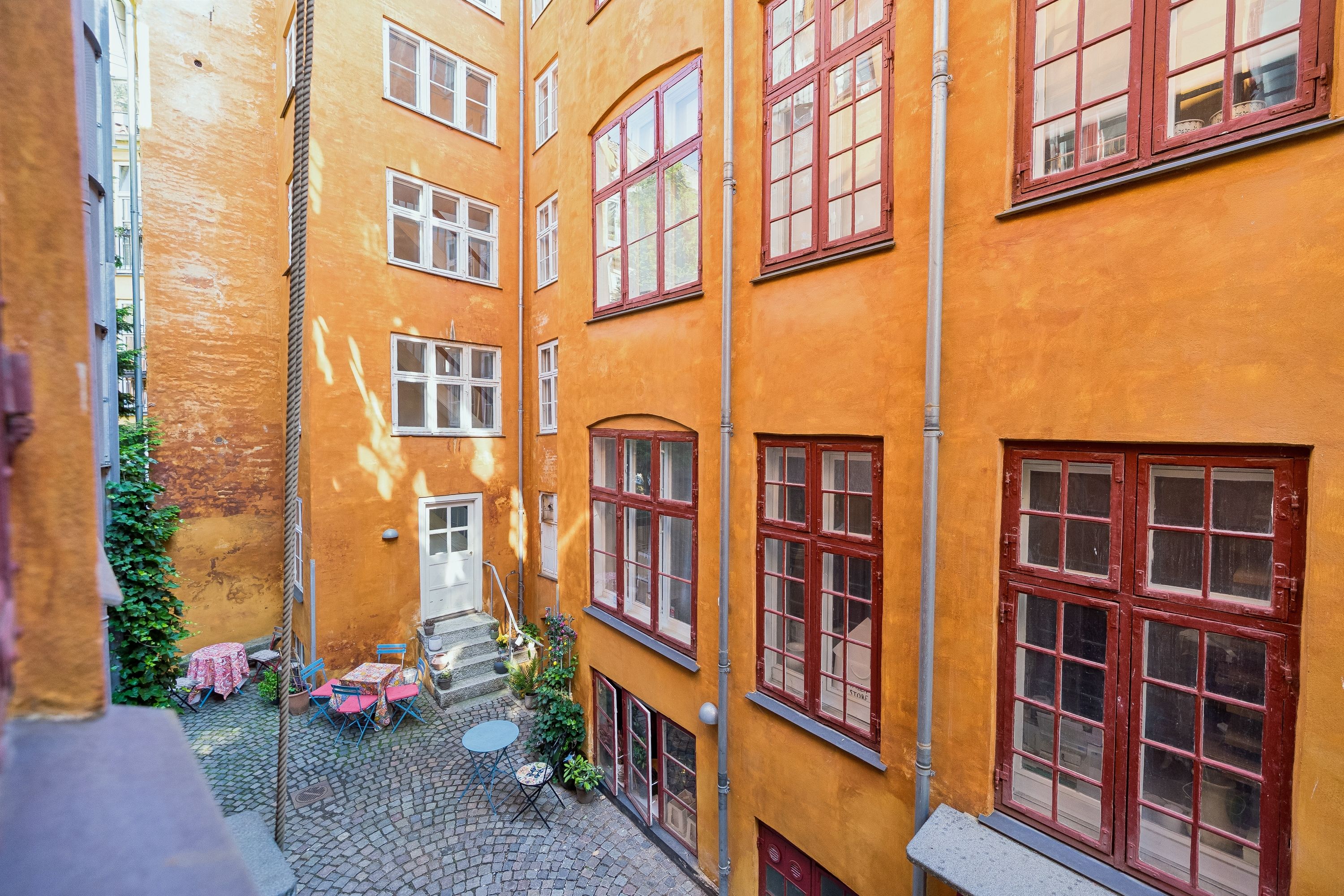 Baghuskontor i historisk bygning med røde ruder og møblerede gårdhave. Vinduesrækker og terrasser med solbelyst gulv og grønne planter.
