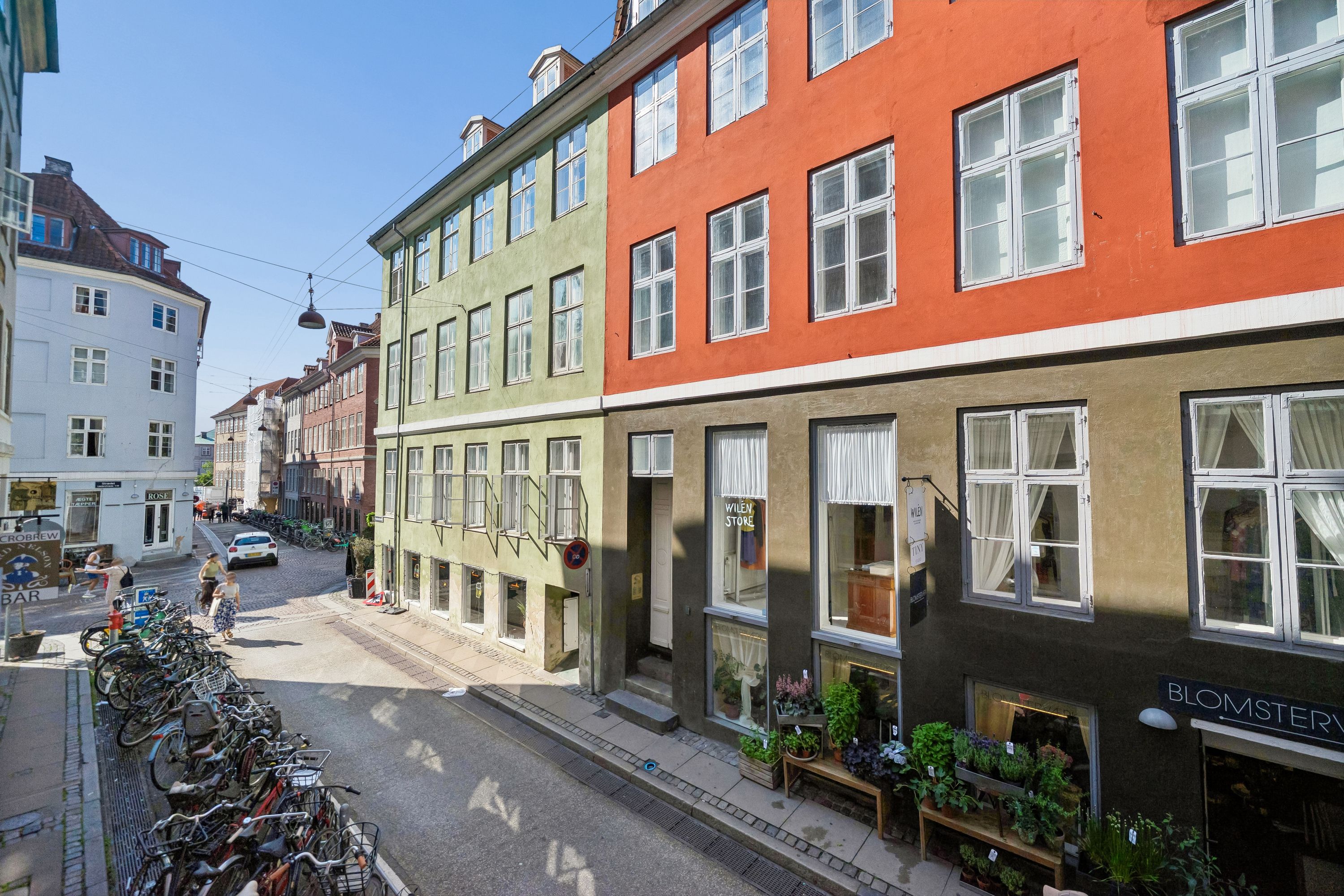 Kontor til leje i København med udsigt til en gammel gade. Røde, grønne og brune bygninger med hvide vinduerne og planter foran. En bil og folk på gaden.