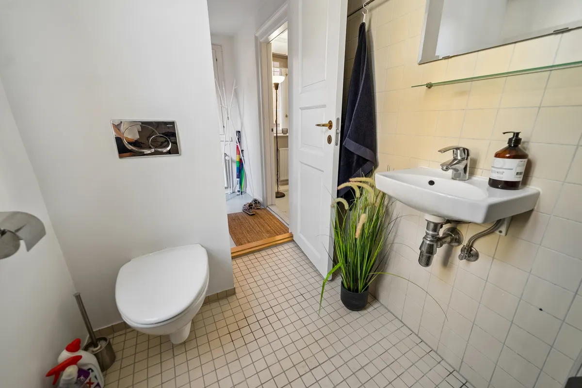 Et lille badeværelse med hvidt toilet, hvidt vask og gulv med hvide og grå firkantede fliser samt lysegule og hvide kakeløg.