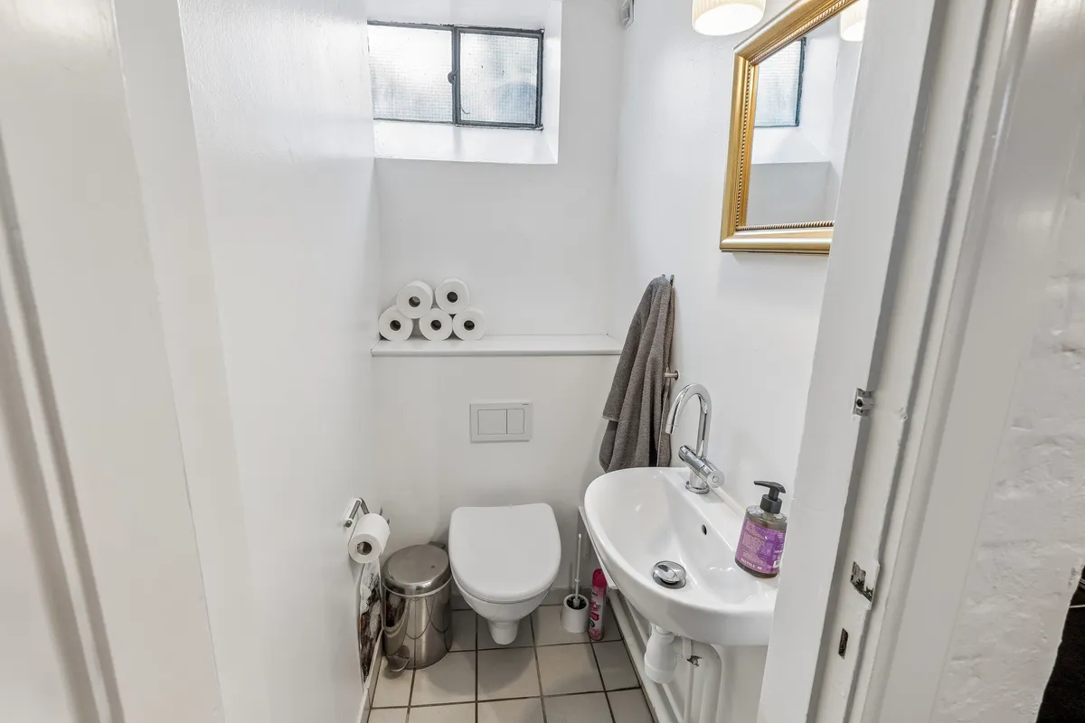 Et lille badeværelse med toilet, håndvask, to toiletpapirruller og et lysekron på væggen, hvor der er et spejl og flere toiletpapirruller