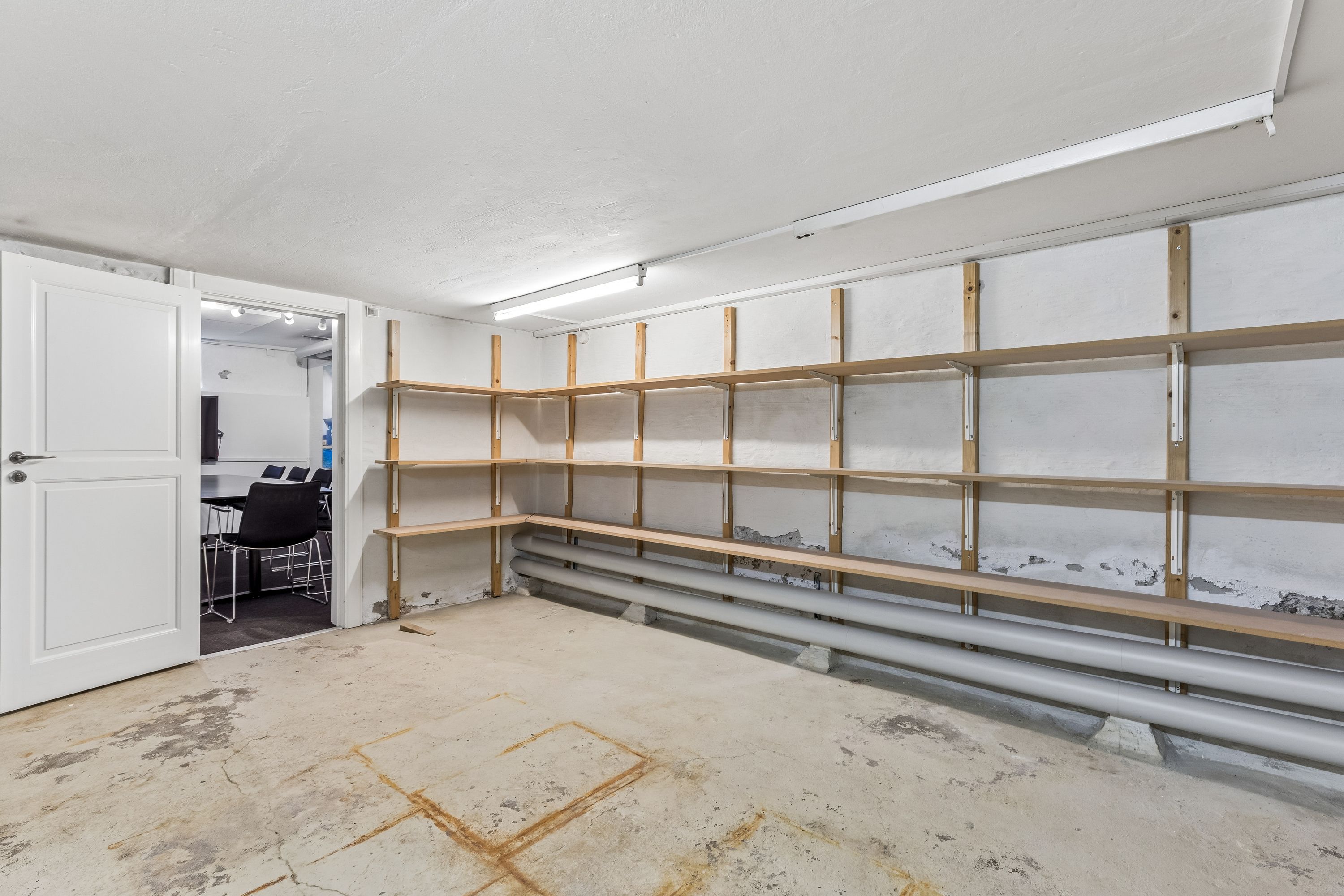 Et tomt lager med lyse gulvvægge og loft, hvor lyse lysekroner er installeret. Der er flere lyse brædder i rummet, som er placeret på væggen. Væggen er malet hvidt og gulvet er beton.