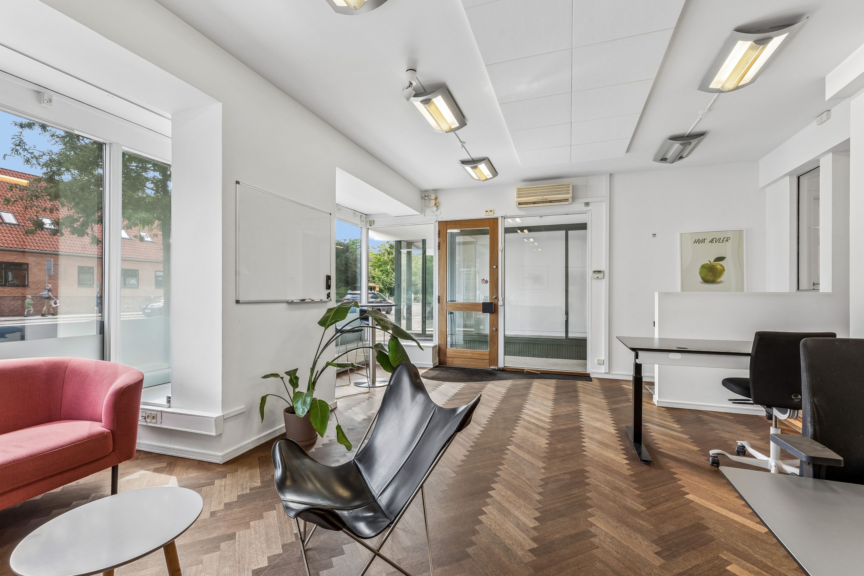 Inde i butik med højt loft, hvide vægge og lyse loftslampe. Sort læderstol og rødt sofa samt skrivebord og kontorstole. Dekorative objekter og plante i køkken.