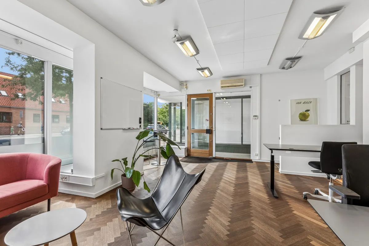 Inde i butik med højt loft, hvide vægge og lyse loftslampe. Sort læderstol og rødt sofa samt skrivebord og kontorstole. Dekorative objekter og plante i køkken.
