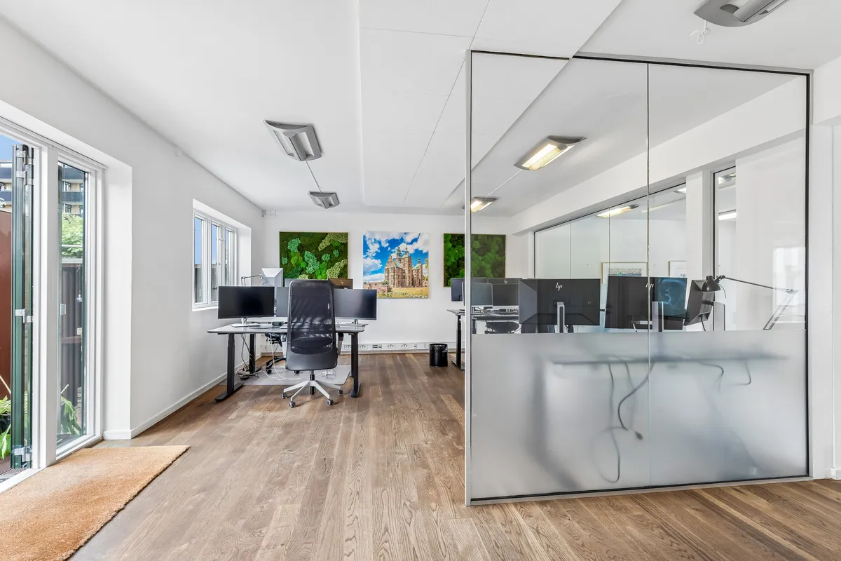 Kontor med lyse møbler, højt loft og store ruder, to store skriveborde med monitorer, og to møbler med stol og plante