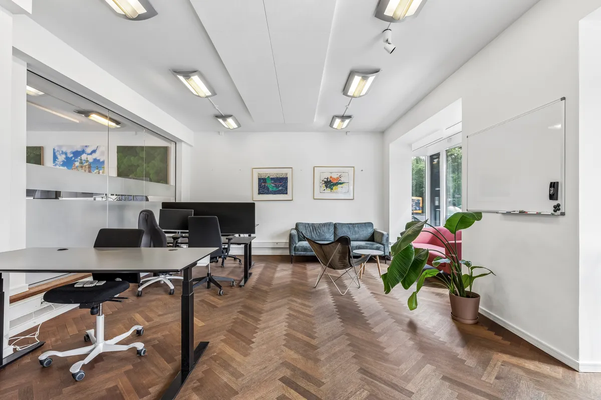 Kontor med skrivebord, stol, sofa, plante og kunstværker i et lyse og moderne kontor med trægulv og hvide vægge.